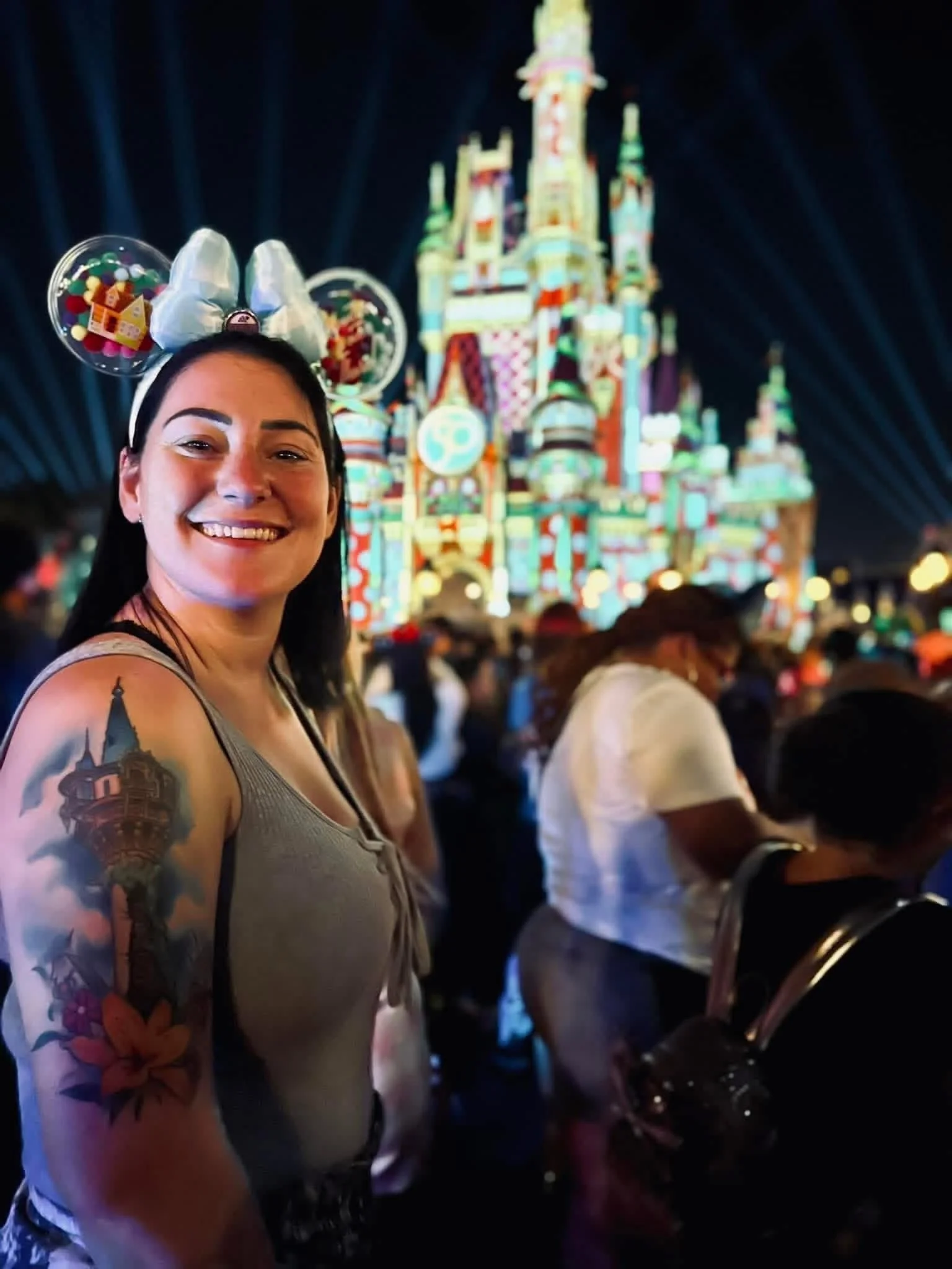 A woman with tattoos on her arm, wearing a headband with Mickey Mouse ears, smiling at a Disney theme park at night with illuminated castle in the background.