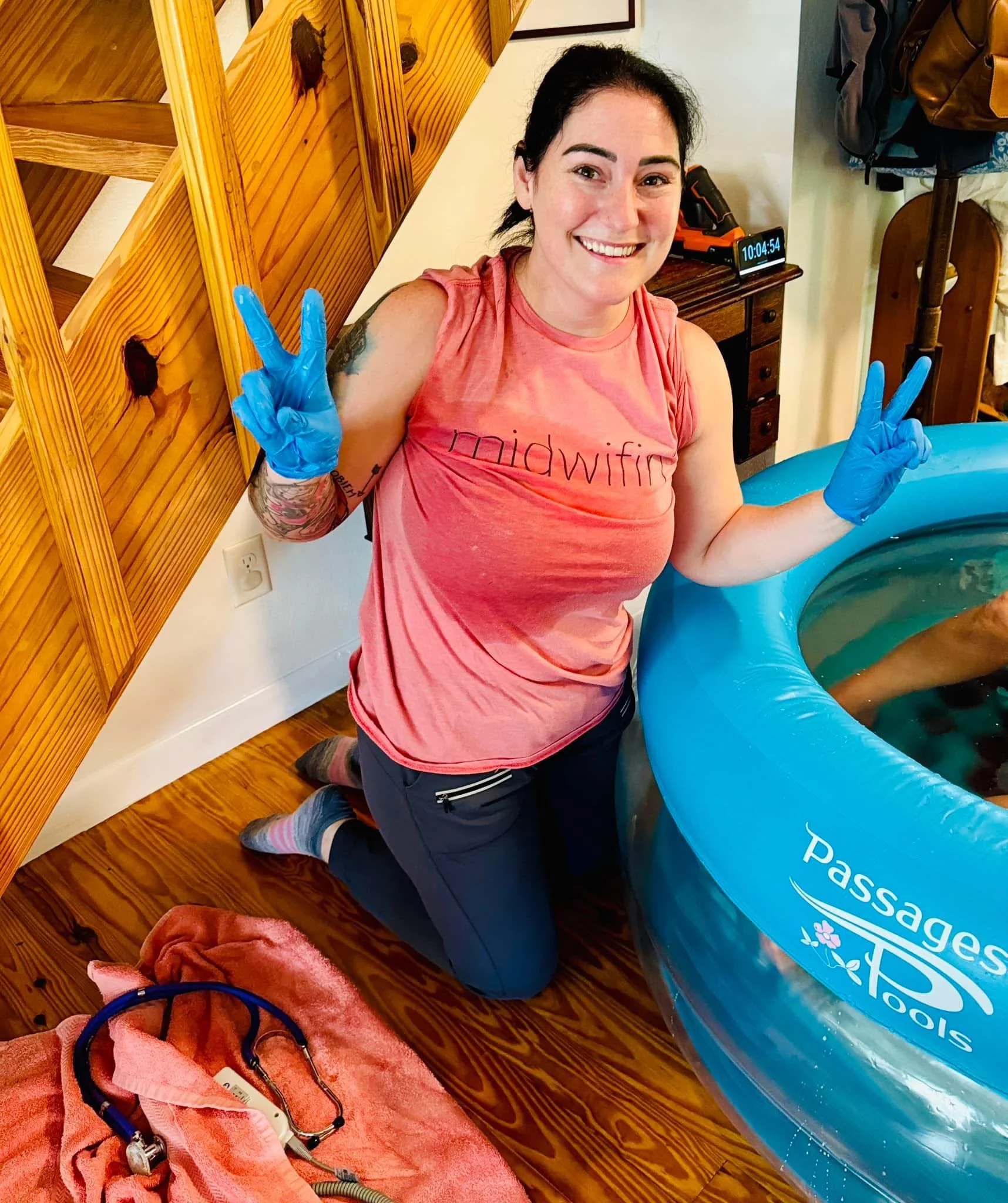 Woman smiling and wearing blue gloves, sitting beside an inflatable pool with a pass tag logo, in a room with wooden furniture and a digital clock showing 10:04.