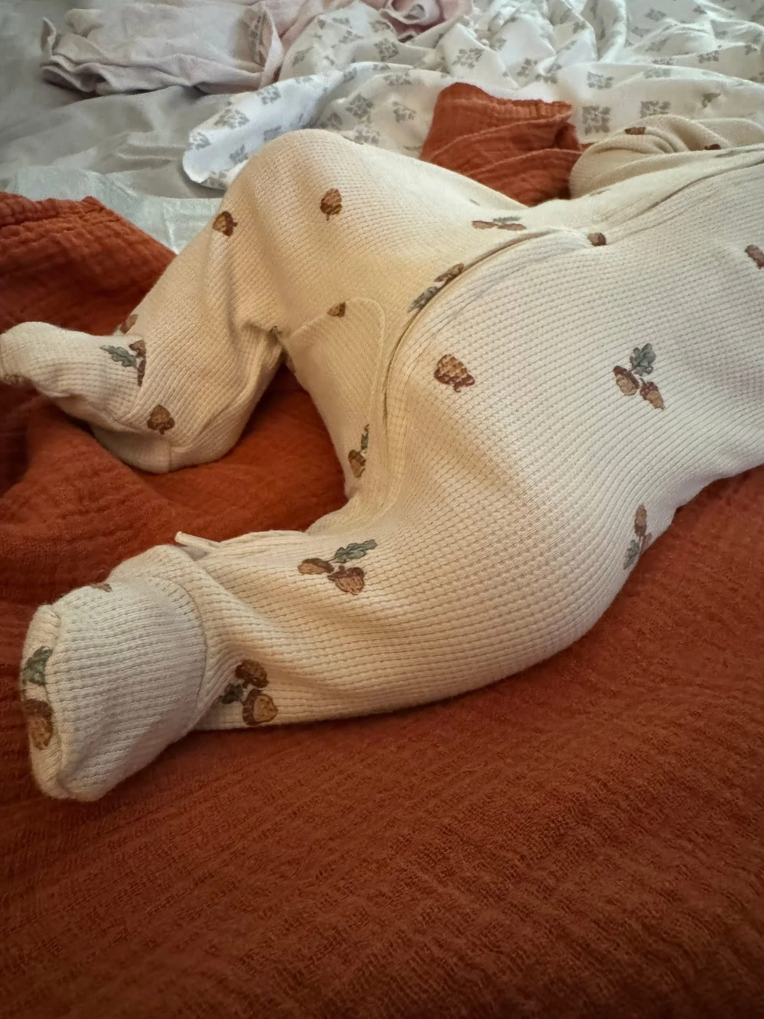 Close-up of a person wearing cream-colored knit pajamas with a small bear pattern, lying on a rust-colored bedspread and surrounded by crumpled blankets.
