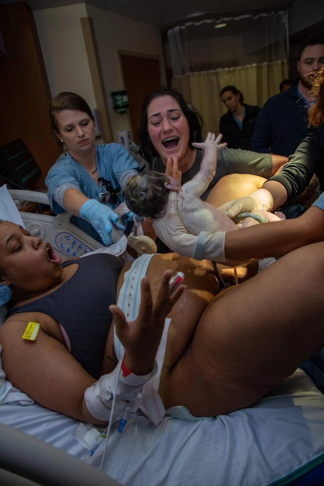 A woman giving birth in a hospital with medical staff assisting. The woman appears to be in the midst of delivery, with her mouth open as if crying or shouting, and her body is in labor. Medical personnel are helping with the delivery, including a woman in scrubs holding the newborn after delivery, who appears to be a healthcare worker.