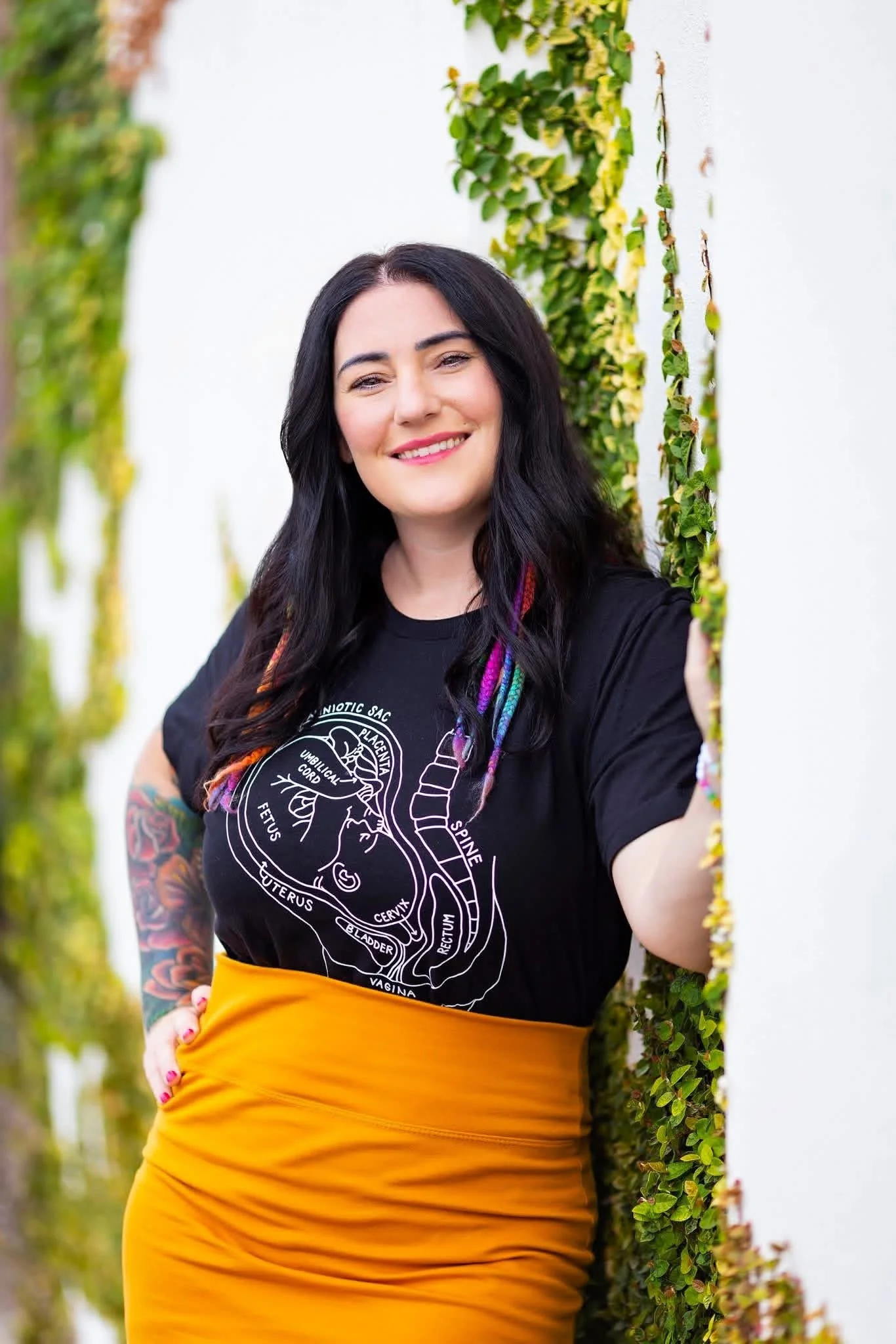 A smiling woman with dark hair and tattoos on her right arm, wearing a black T-shirt with a diagram of the human brain and a mustard yellow skirt, standing near a white wall with green and yellow climbing plants.