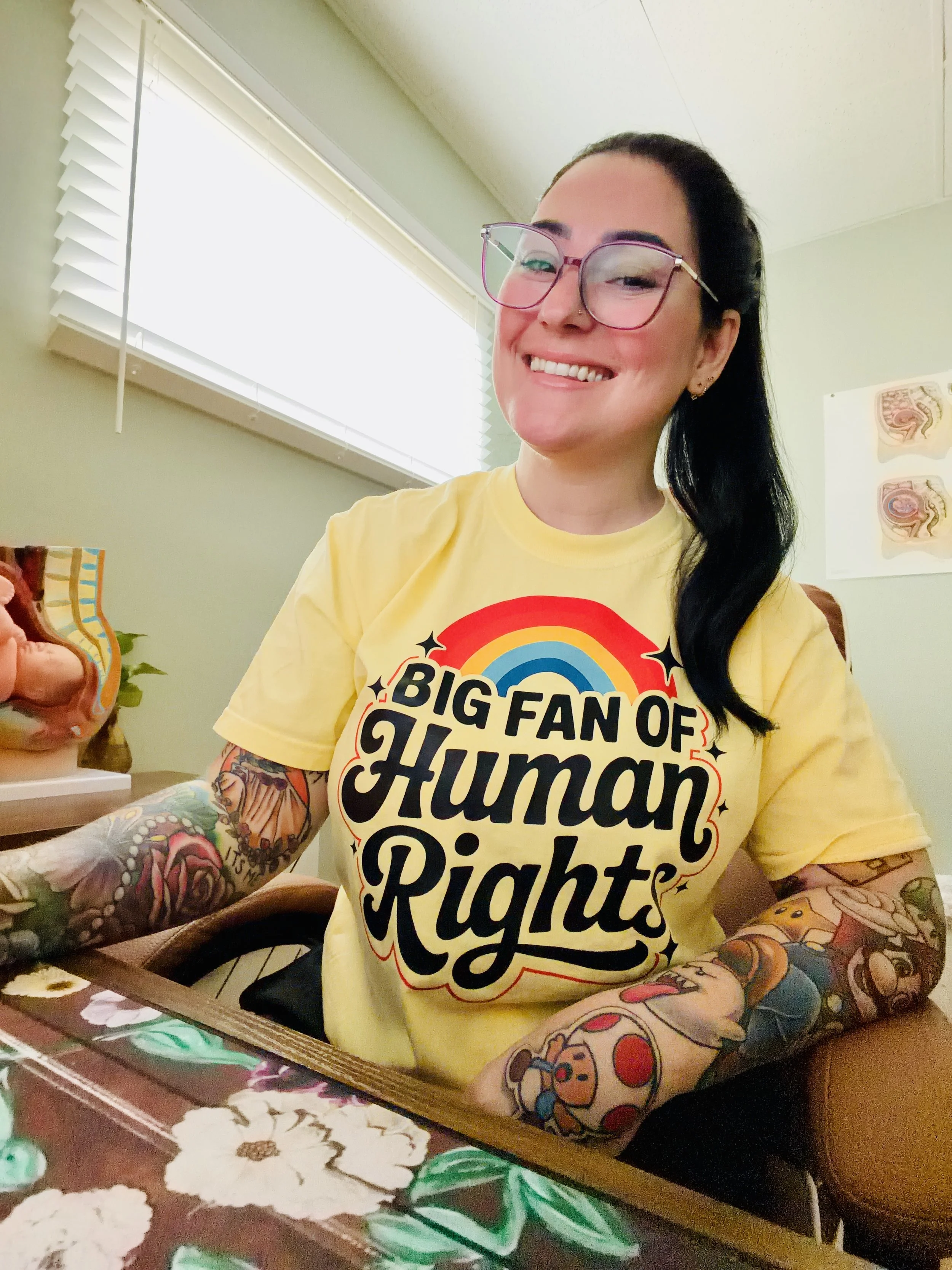 A woman with long black hair, glasses, and tattoos on her arms, sitting at a table, wearing a yellow T-shirt with the text "Big Fan of Humble Rights" and smiling at the camera. There are blinds on a window behind her, and a poster with anatomical sketches on the wall.