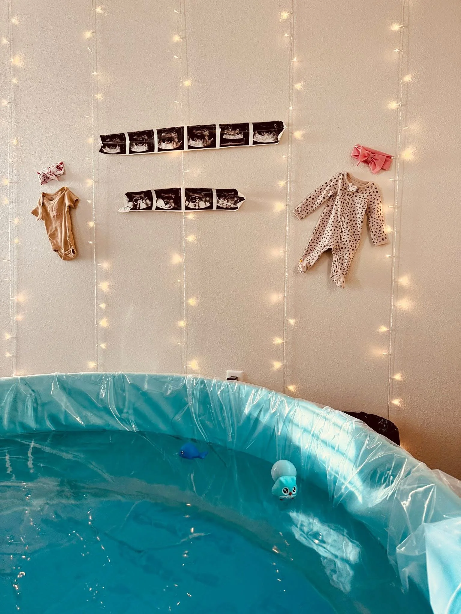 Indoor scene with a small swimming pool or water area, decorated with string lights on the wall. The wall features several ultrasound images and baby clothes hanging, including a pink bow, a beige onesie, and a polka-dotted outfit.