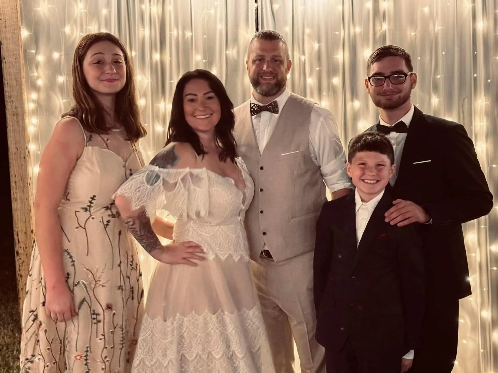 A group of five people, including a bride in a white lace wedding dress, standing in front of a backdrop decorated with lights, at a wedding celebration.