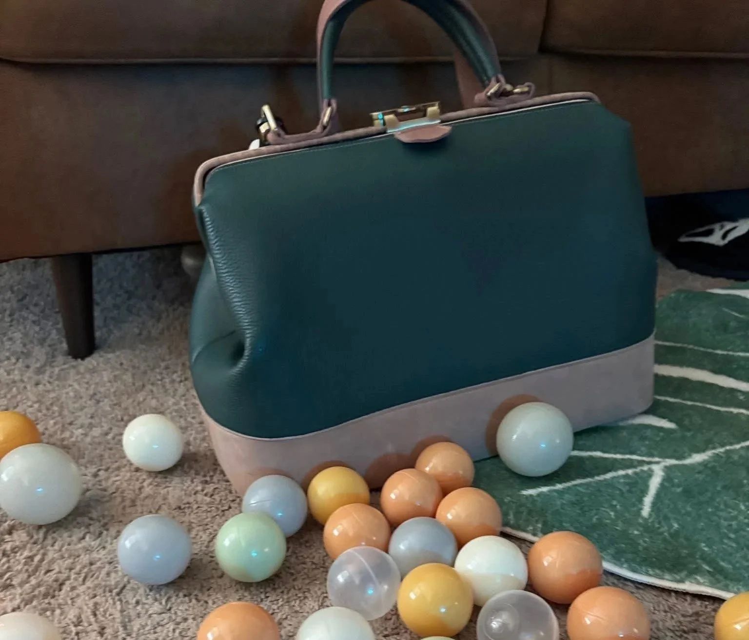 A green and gray vintage-style briefcase sitting on a carpeted floor amidst plastic balls of various colors, with a dark wooden piece of furniture in the background.