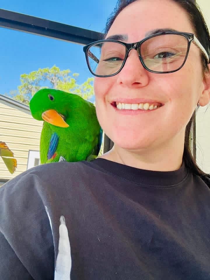 Woman smiling with a vibrant green and blue parrot perched on her shoulder outdoors, with a house, trees, and a clear blue sky in the background.