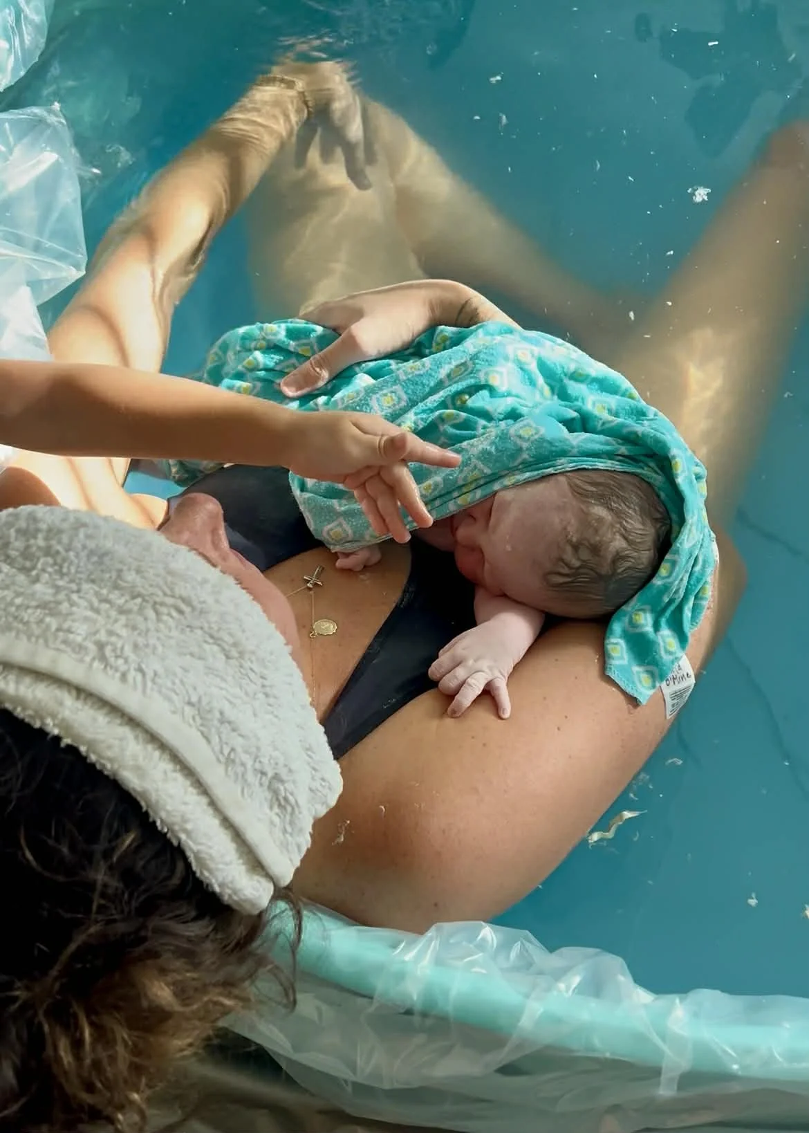 A newborn baby being held on a woman’s chest after birth in a water birth, with a turquoise towel covering the baby's head and water surrounding them.