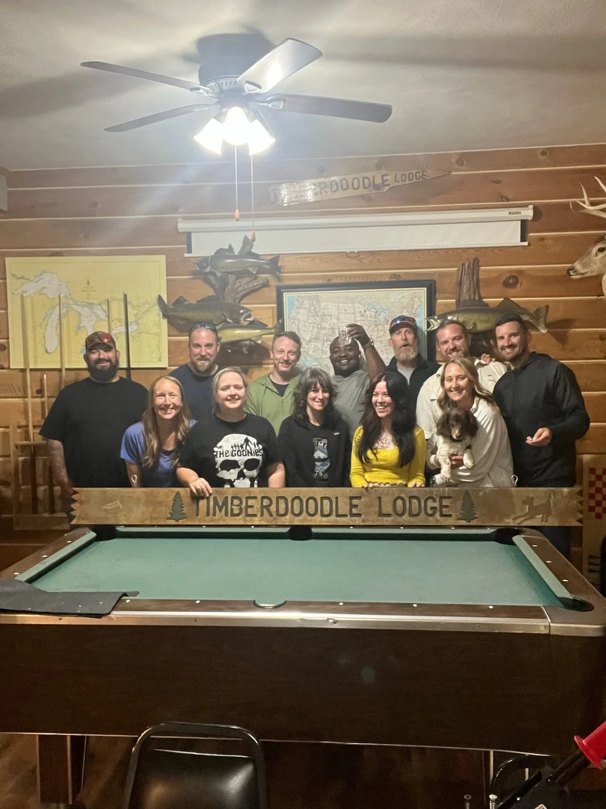Group of people smiling and gathered in a rustic wooden room with fishing decor, standing behind a pool table with a Timberdoodle Lodge sign, some raising drinks, with a dog and mounted fish on the wall.