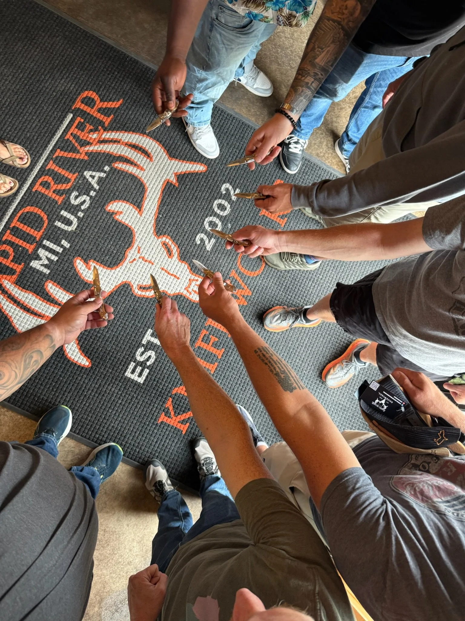 People standing in a semicircle on a floor mat with a logo, holding small knives.