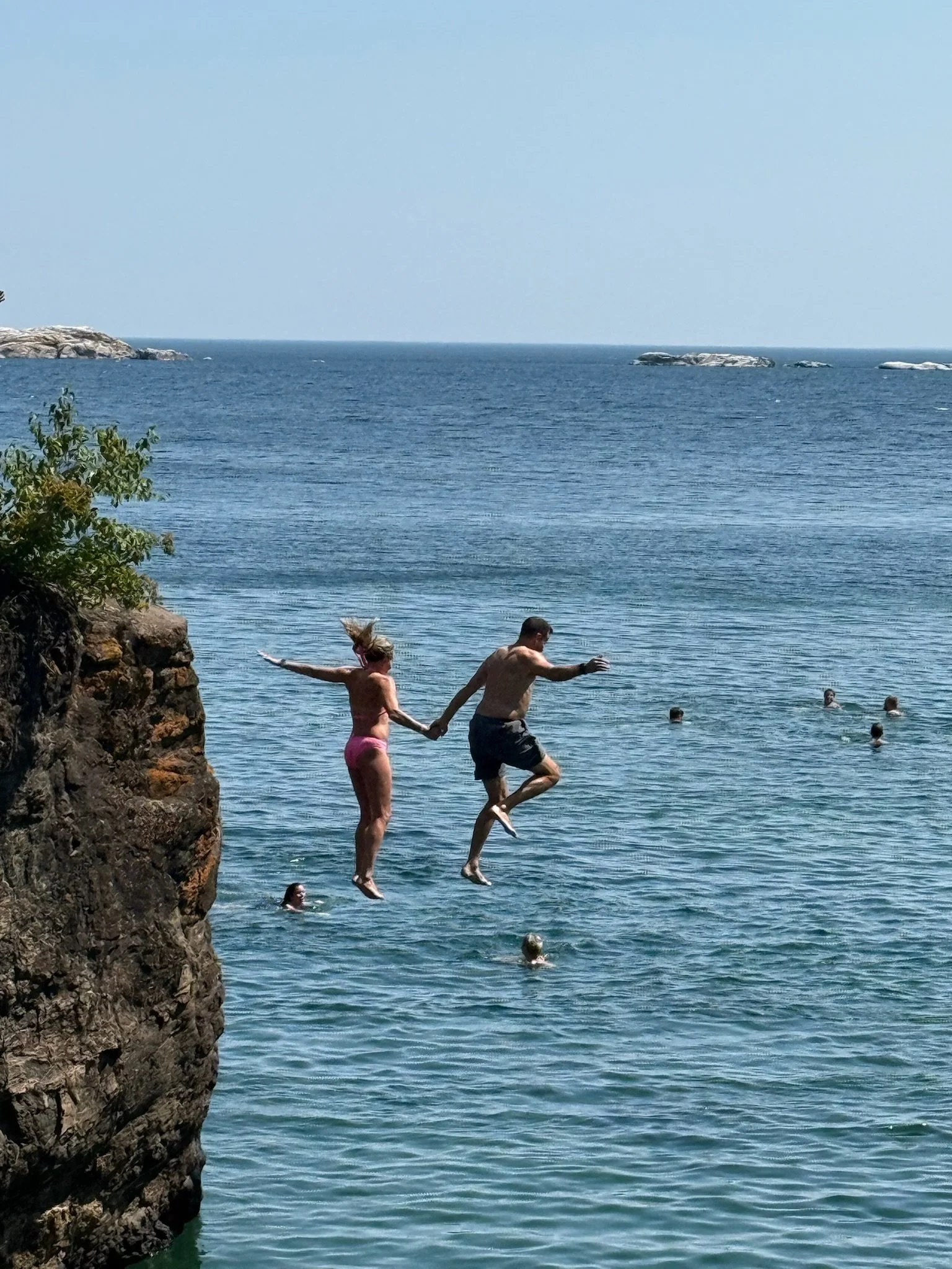 People jumping off a rocky cliff into a lake near the ocean on a sunny day, with others swimming in the water and small islands in the distance.