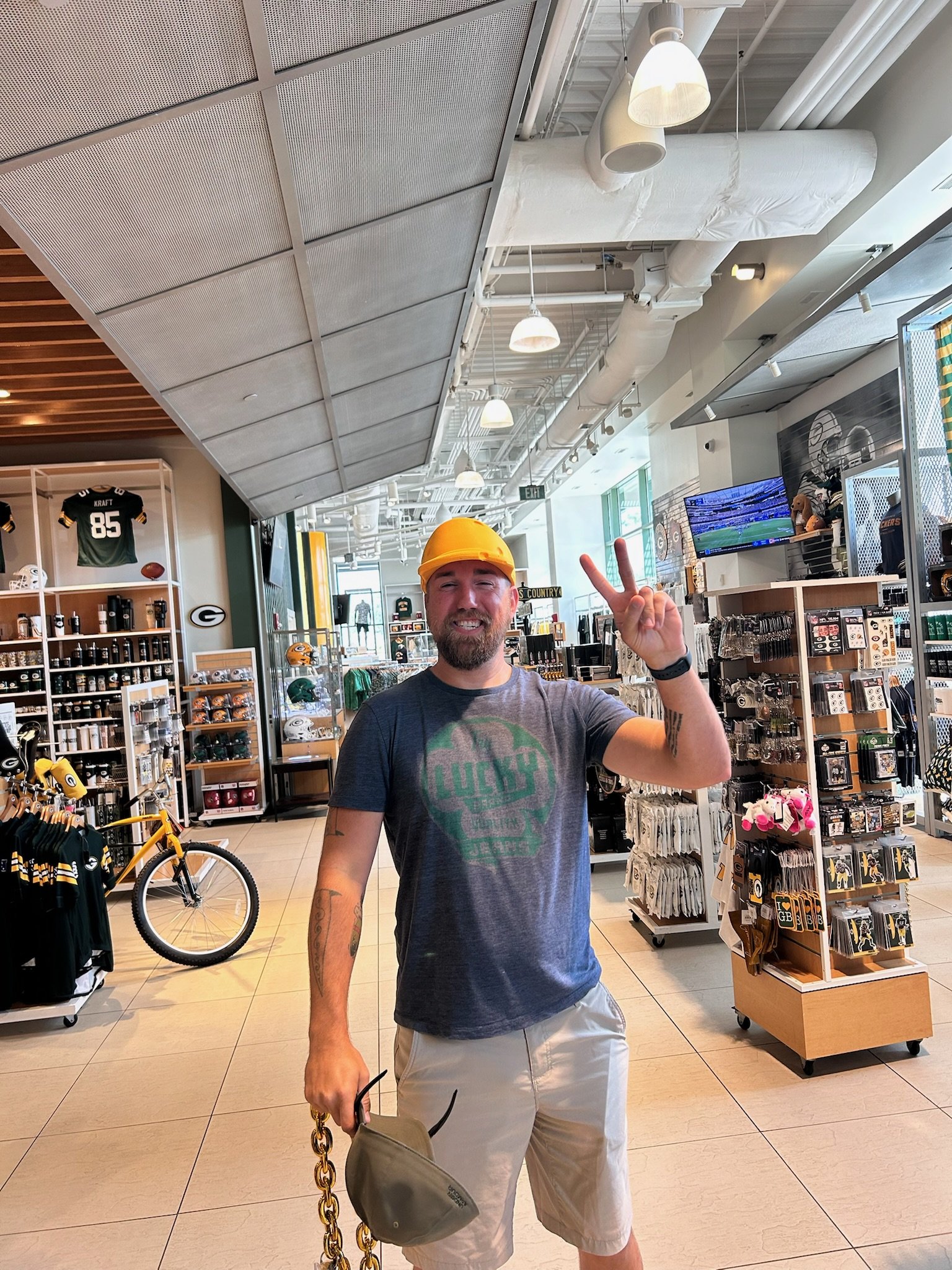 A man in a yellow construction helmet smiling and flashing a peace sign inside a sports merchandise store. The store has sports jerseys, helmets, and other memorabilia on display, and features a modern industrial design with exposed ducts and hanging lights.