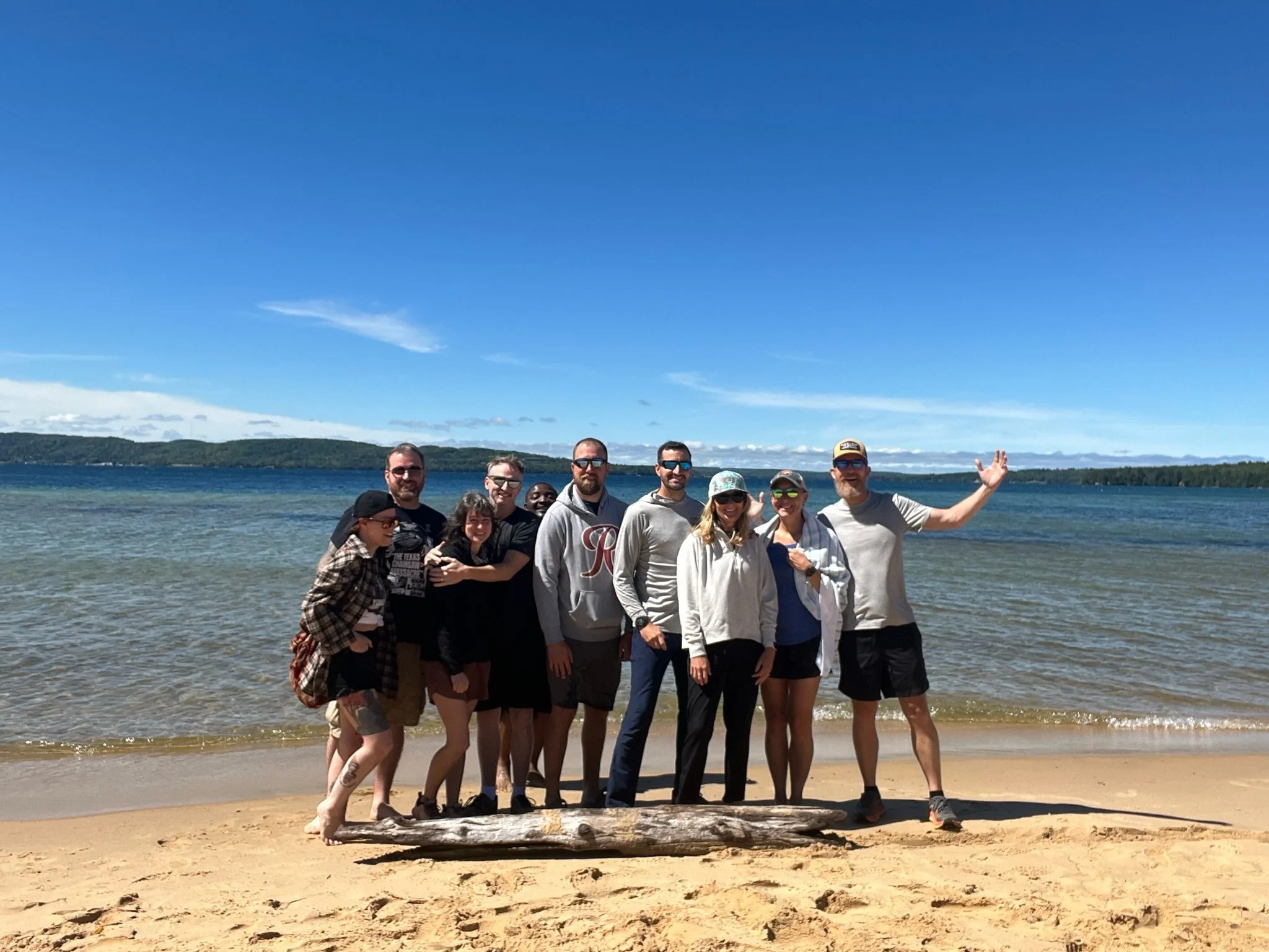 Group of ten people standing on a sandy beach near the water, with a blue sky and distant hills in the background. They are smiling, some wearing sunglasses and casual clothing, and one person is waving.