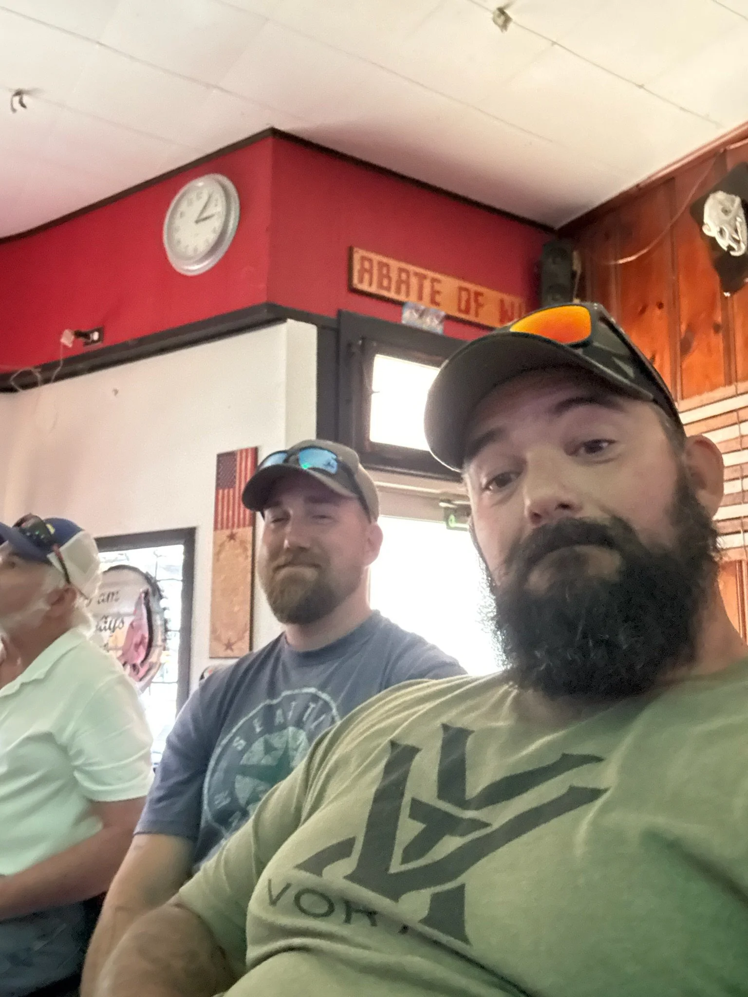 Three men sitting indoors, one with a beard wearing a yellow shirt with zigzag pattern, one with a goatee wearing a dark T-shirt, and one in a light-colored shirt with glasses and a cap. Part of a bar or restaurant interior with a clock on the red wall, a sign that reads 'ABATE OF M' partially visible, and wooden paneling.