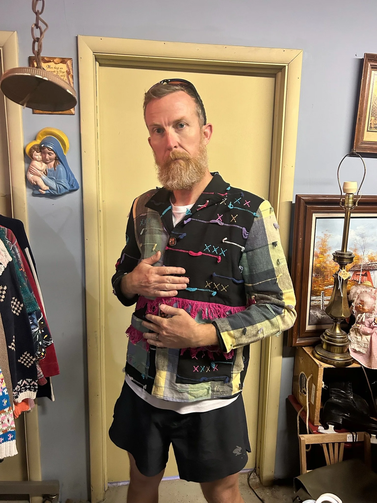 A man with a beard and short hair, wearing sunglasses on his head, a colorful embroidered vest, and black shorts, standing in a room with decorative items on the wall and a lamp.