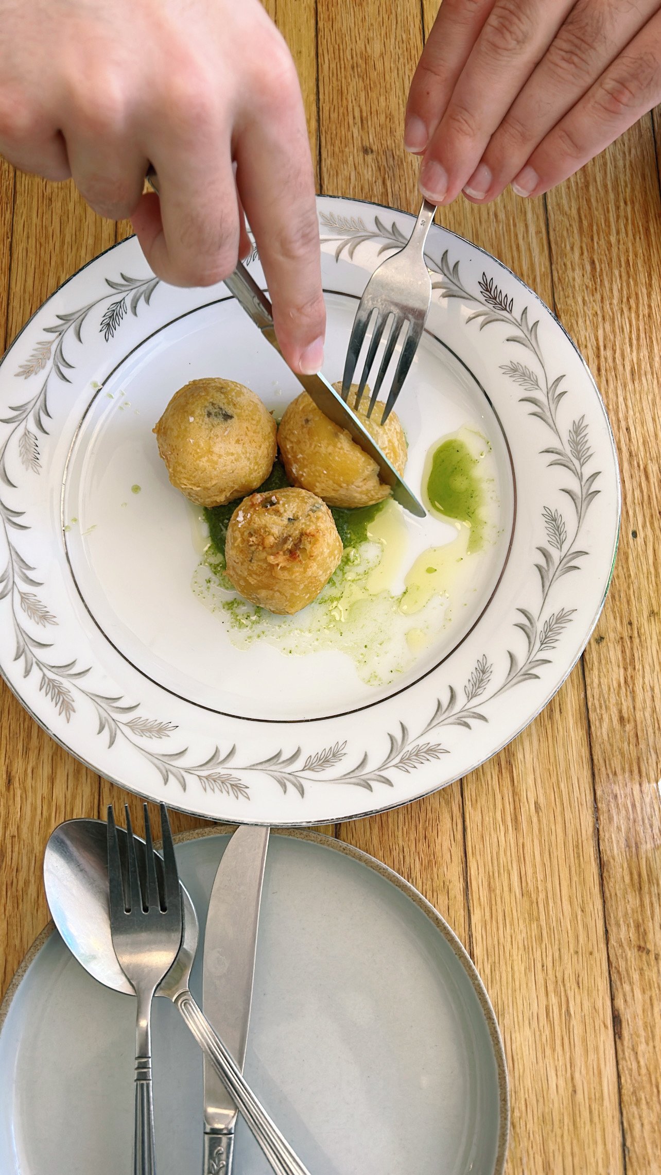 A white plate with a decorative border holding three fried food balls with a green sauce underneath, with hands cutting one of the food balls with a knife and fork. An empty blue plate with forks and knives is in the foreground on a wooden table.