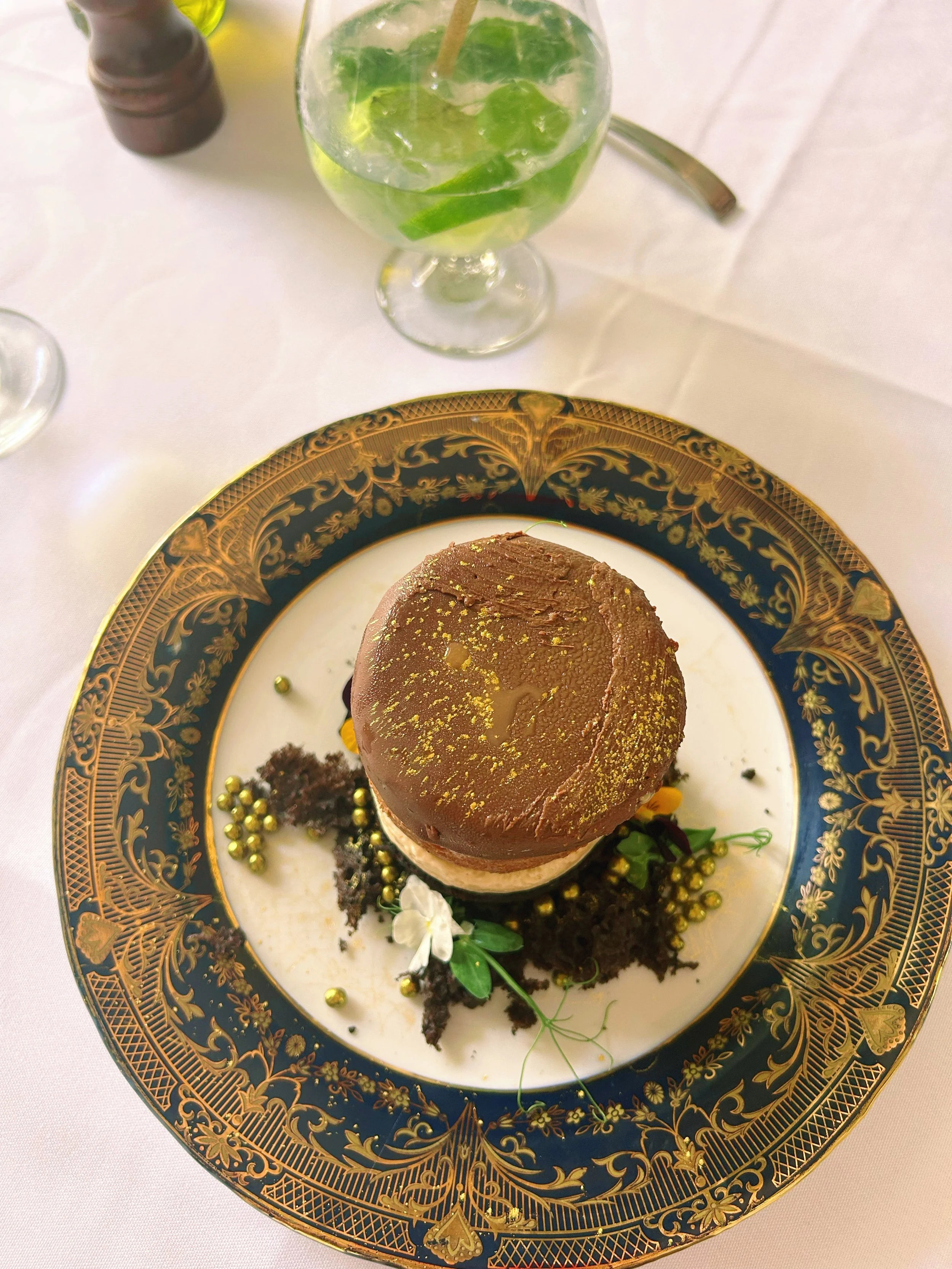 A decorative plate with an ornate gold and dark blue pattern holds a chocolate dessert topped with gold dust and surrounded by chocolate crumbs, small edible gold spheres, green leaves, and flowers. A glass of lime and mint beverage with ice is on the table.