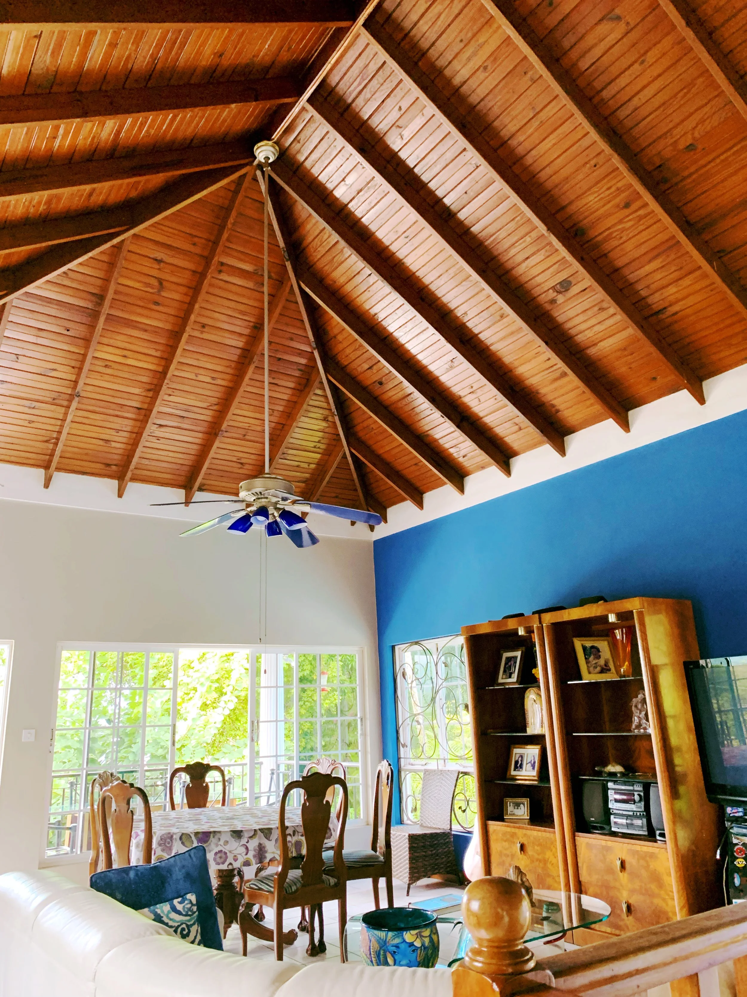 Living room with a wooden vaulted ceiling, ceiling fan, large window letting in sunlight, dining table with chairs, and a blue accent wall with a wooden cabinet.