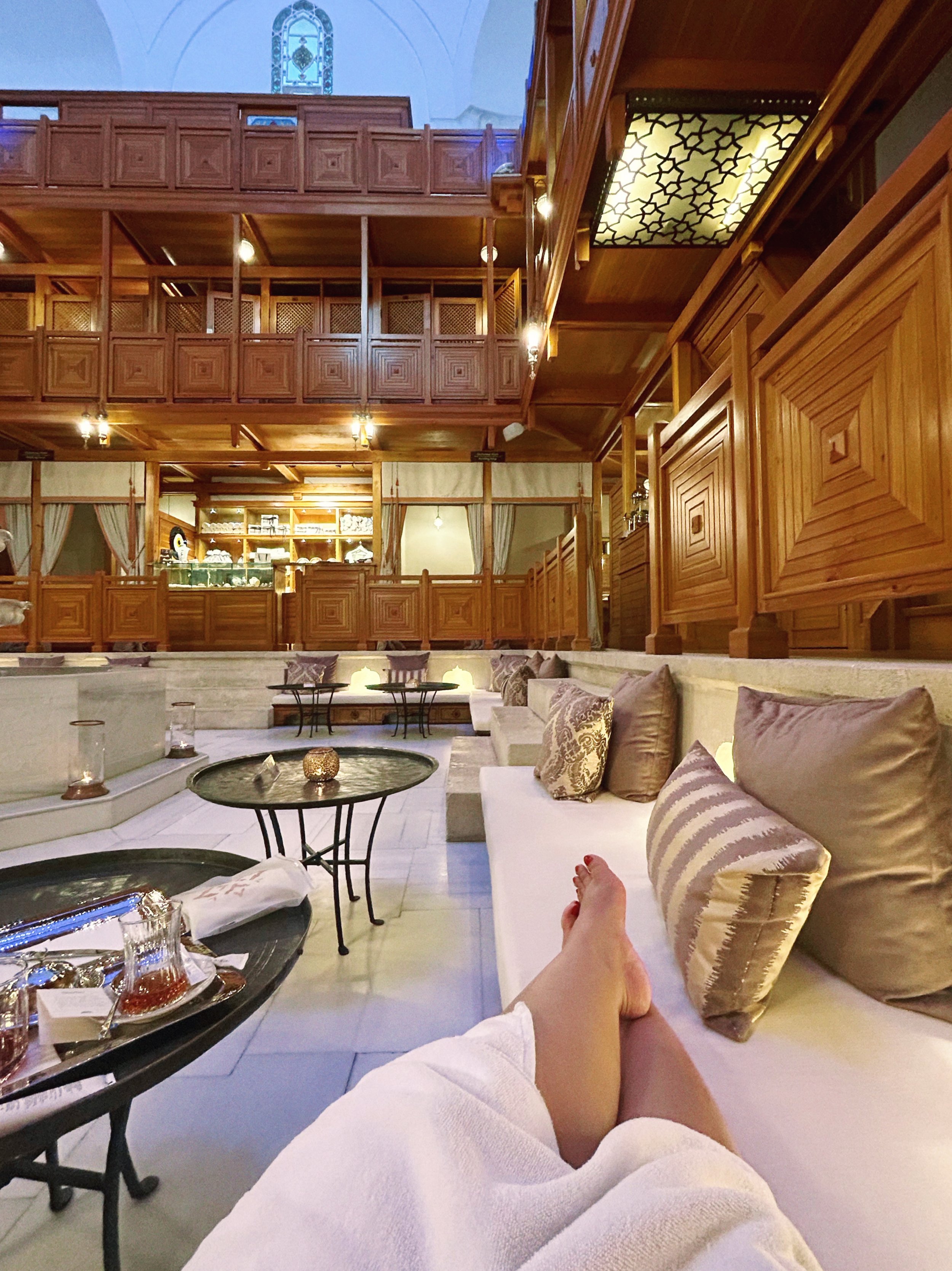 Interior of a cozy lounge area with beige cushioned seating and pillows, small tables with decorative items, and an upper balcony with wooden railing, warm lighting, and a chandelier with a geometric pattern.