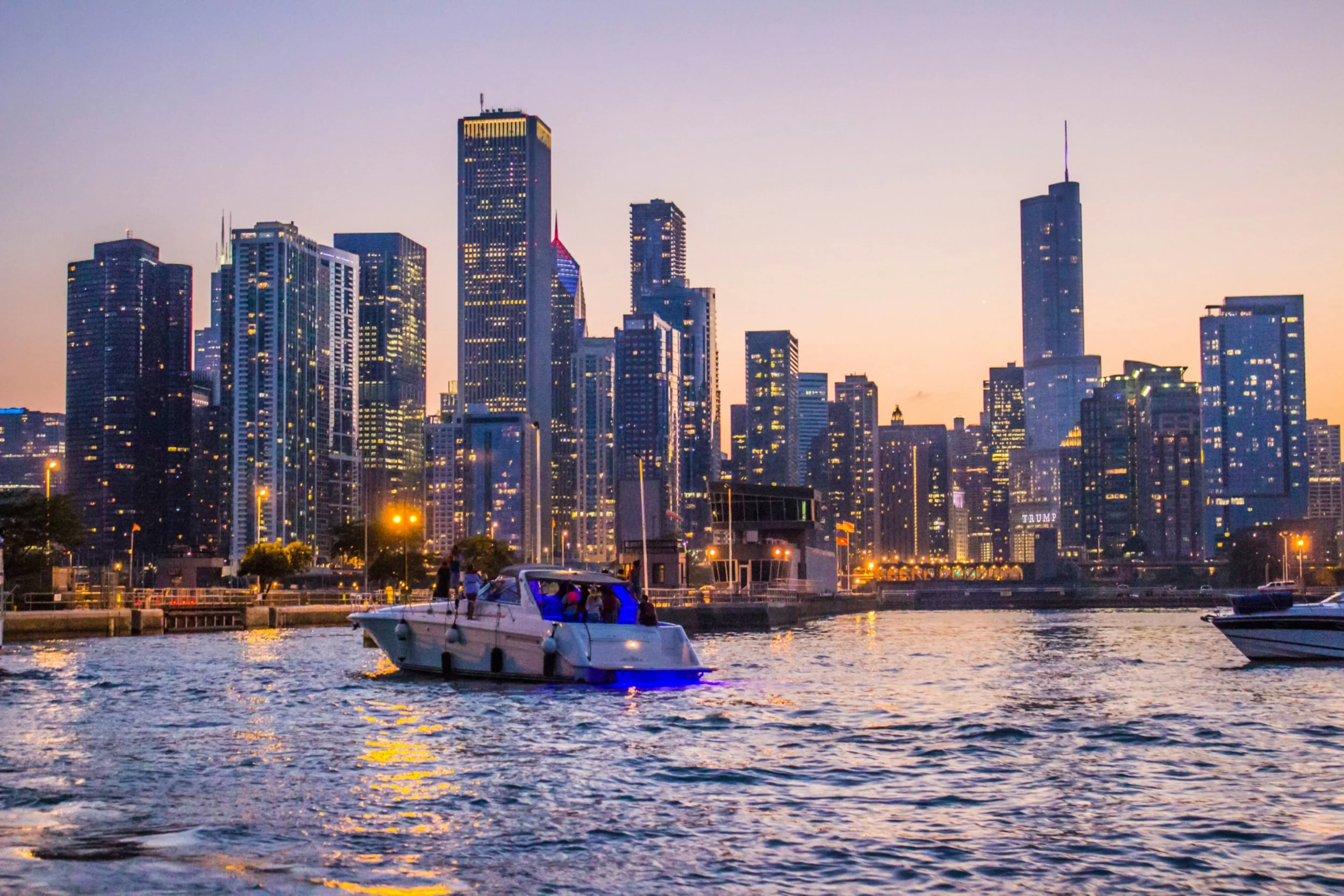 A city skyline with tall buildings illuminated at dusk, viewed across a river with boats and a marina in the foreground.