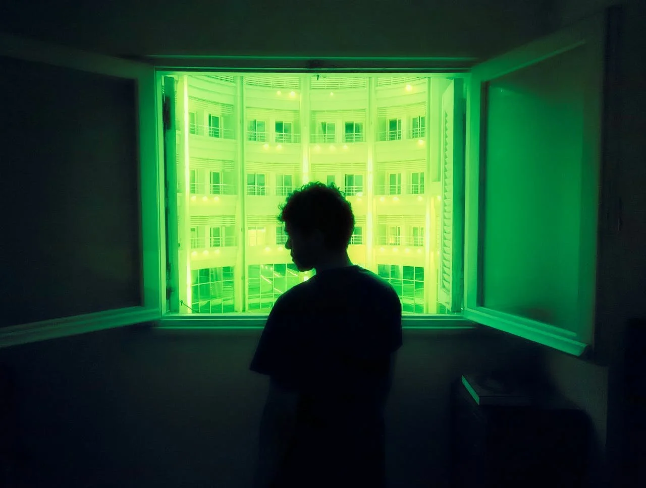 A person standing indoors in front of a window with green neon lights illuminating the building outside.