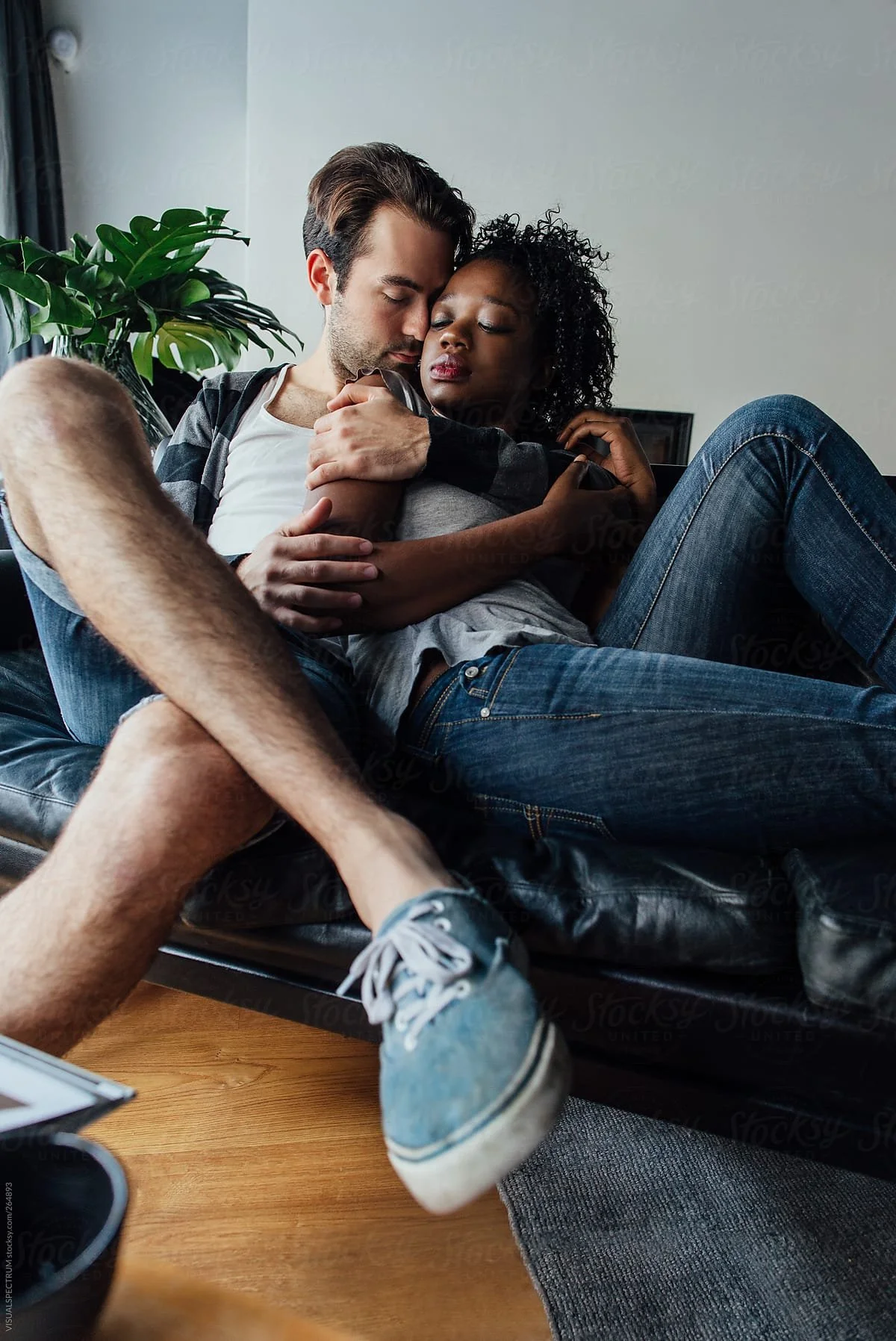 A young man and woman sitting closely on a black leather couch, embracing with eyes closed, in a cozy indoor setting with a plant and a white wall in the background.