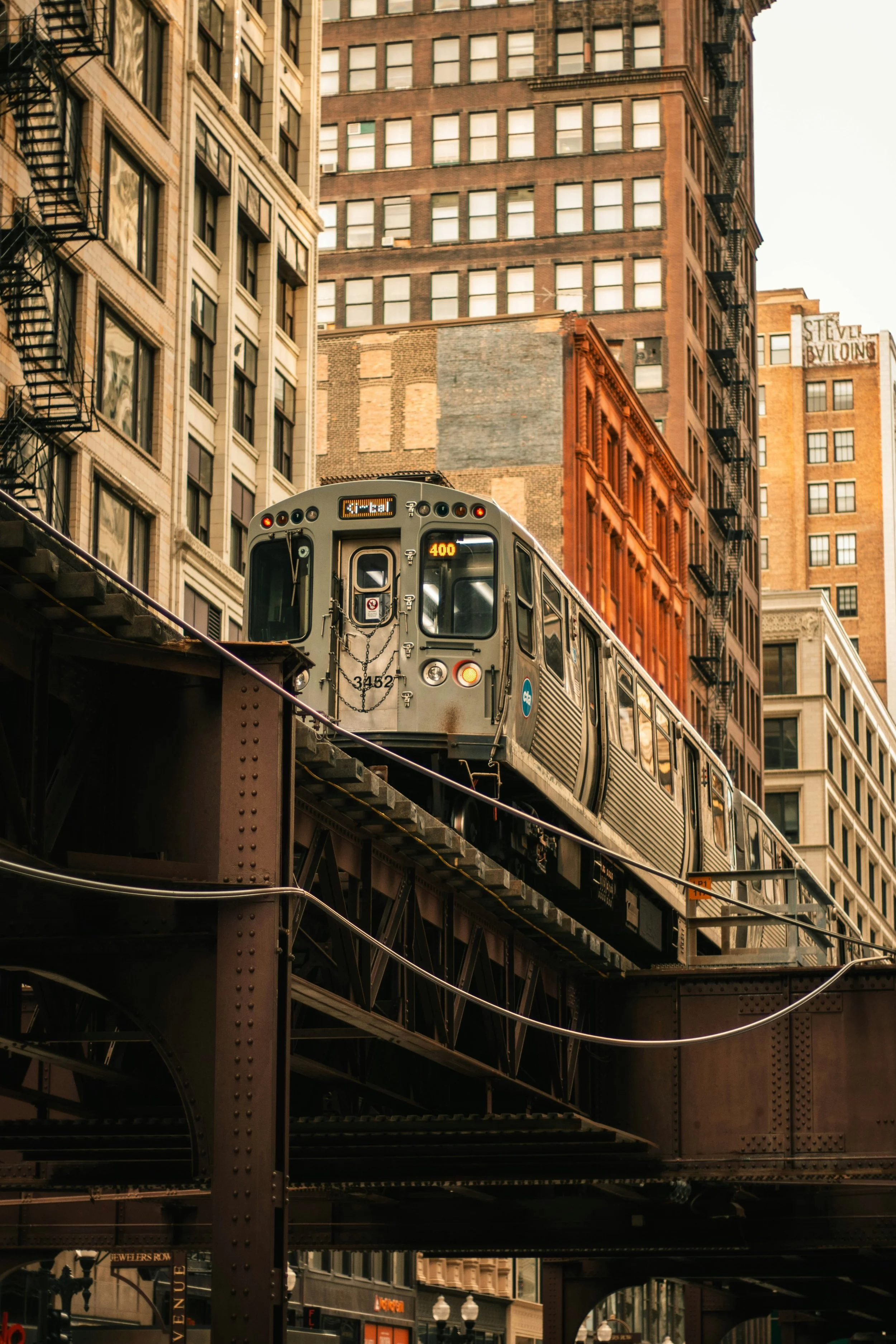 An elevated train moving on an iron track in an urban city surrounded by tall buildings with fire escapes. The background shows a clear sky and multi-story brick buildings.