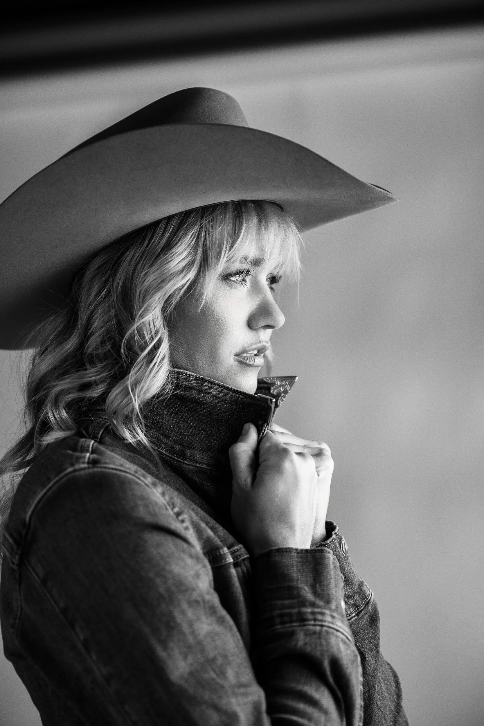 A black-and-white studio portrait of a woman in a wide-brimmed cowboy hat and denim jacket, looking to the side.