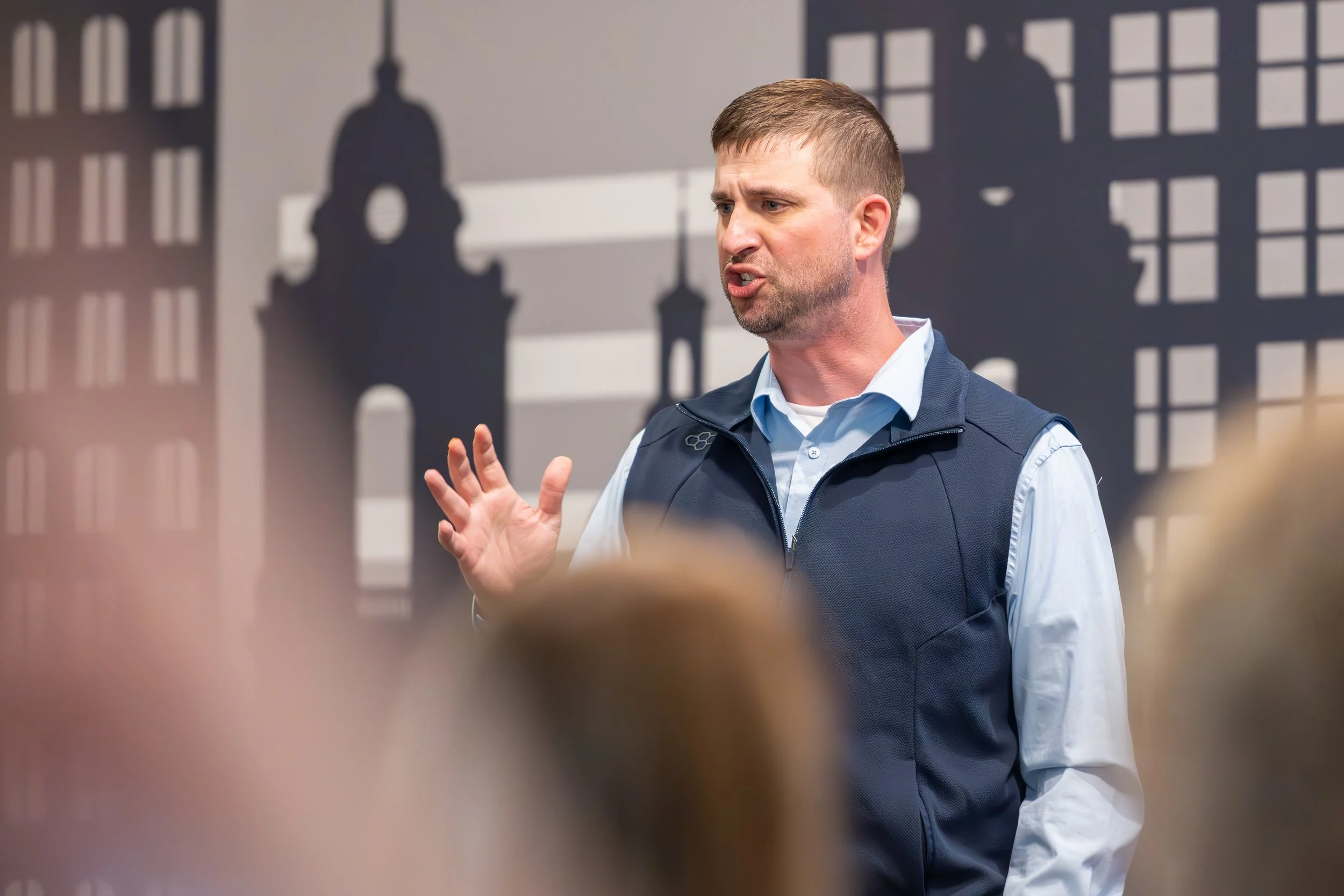 Keynote speaker in vest speaking to an audience against a city skyline silhouette backdrop
