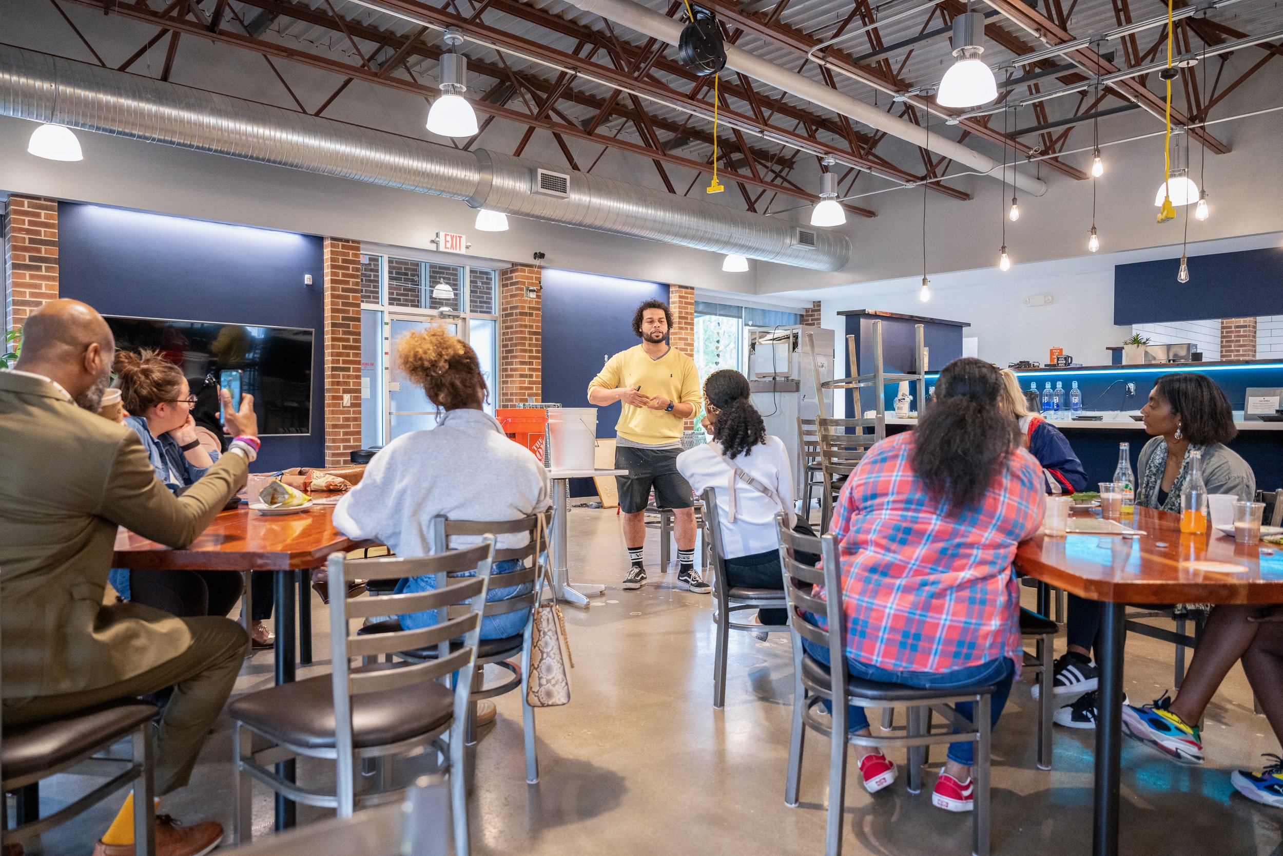 Cafe owner in yellow shirt presenting to a seated group inside a casual restaurant space