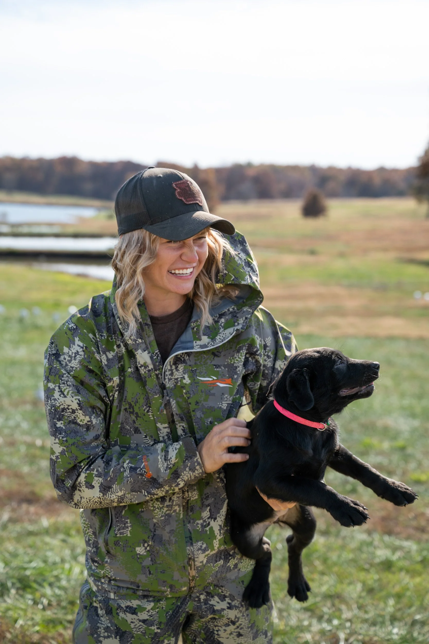 A smiling trainer in camo holds a black Labrador puppy in a green field.
