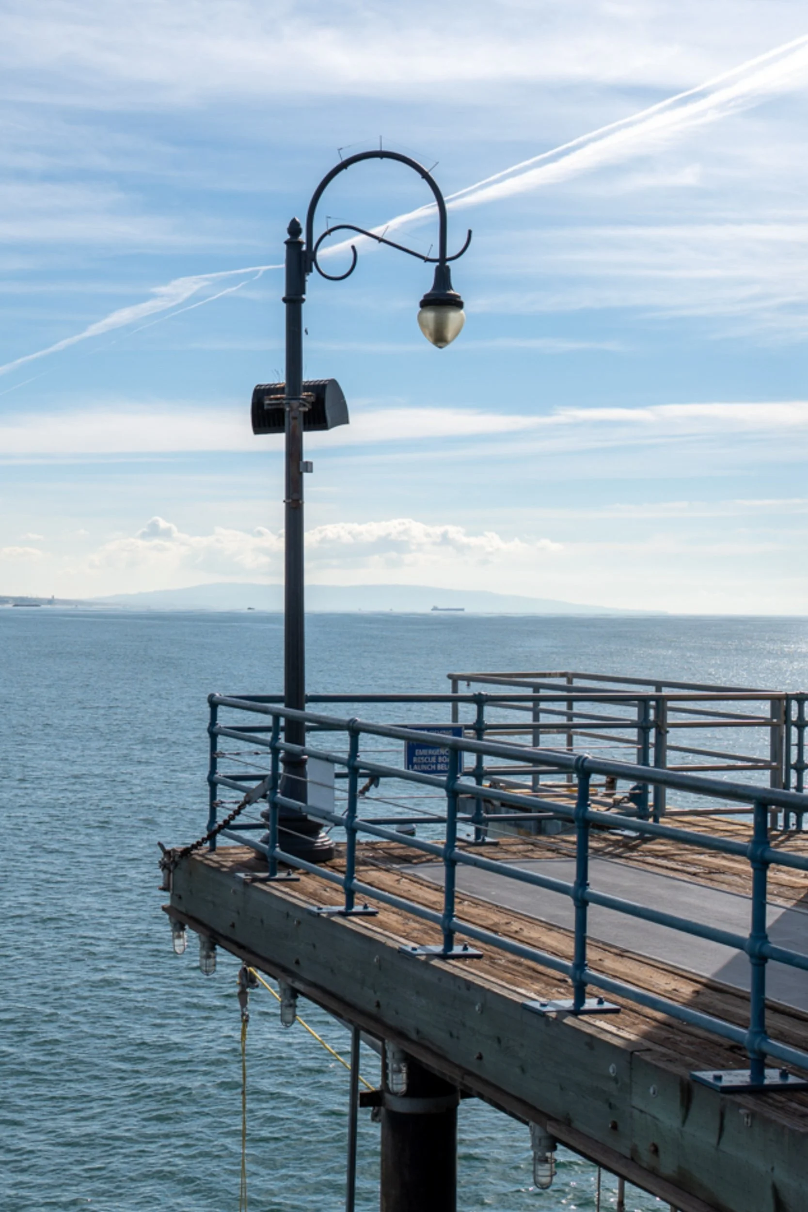 Santa Monica pier extending over the Pacific Ocean, vintage lamp post in the foreground, mountains faint in the distance.
