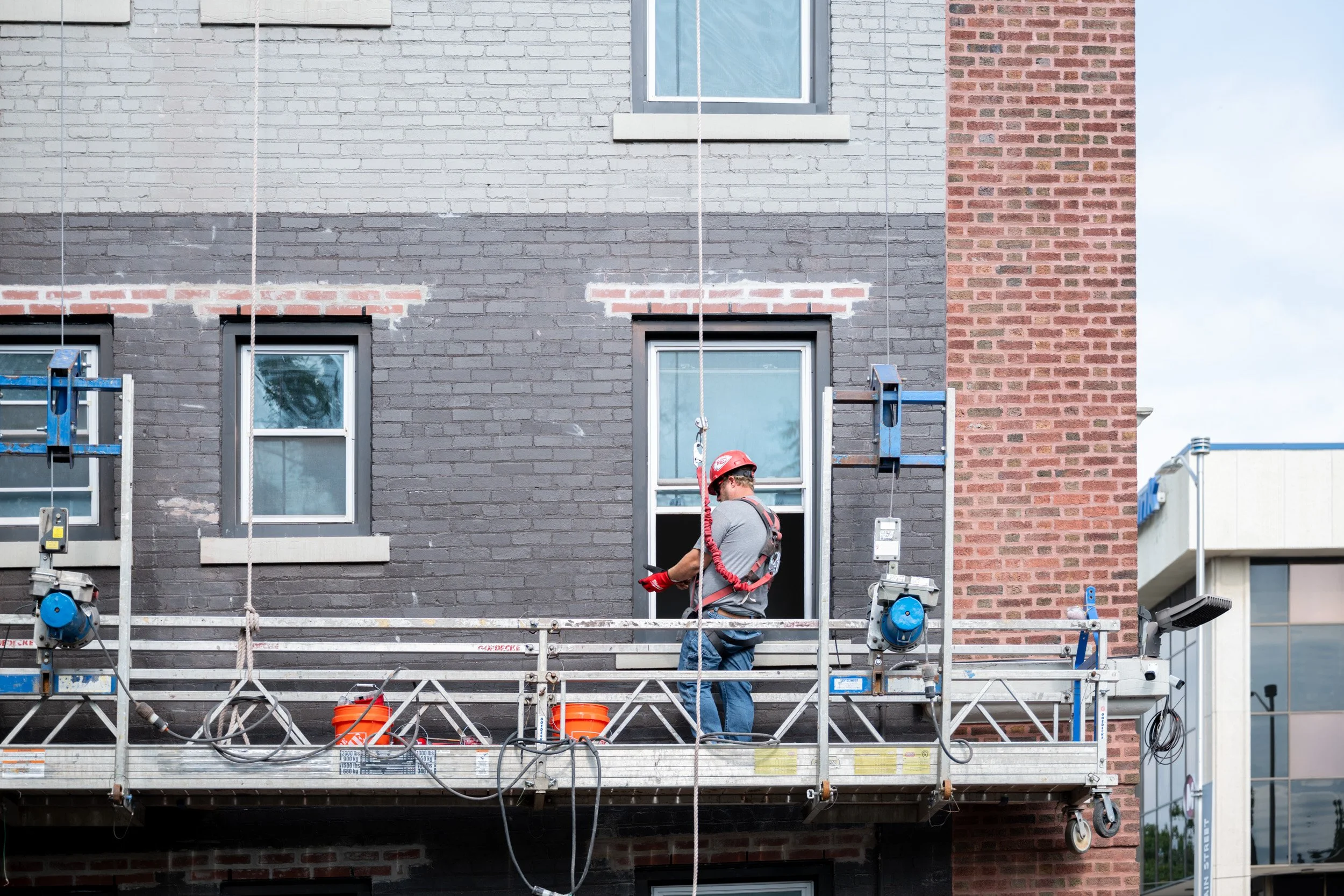 Construction worker in hard hat and harness on a suspended scaffold against a brick facade