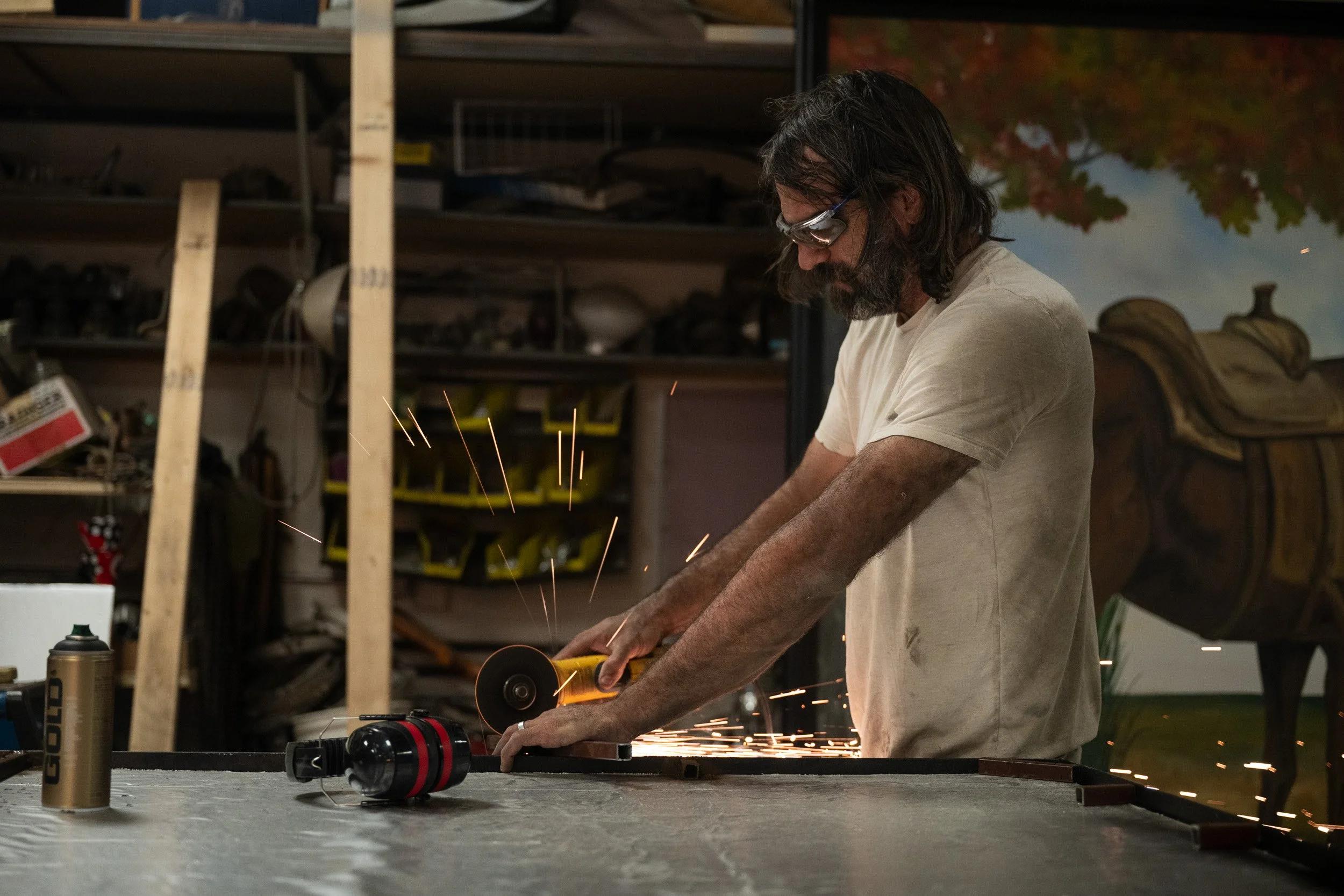 Metal artist grinding a steel frame, sparks flying, beside one of his hand-painted pastoral canvases