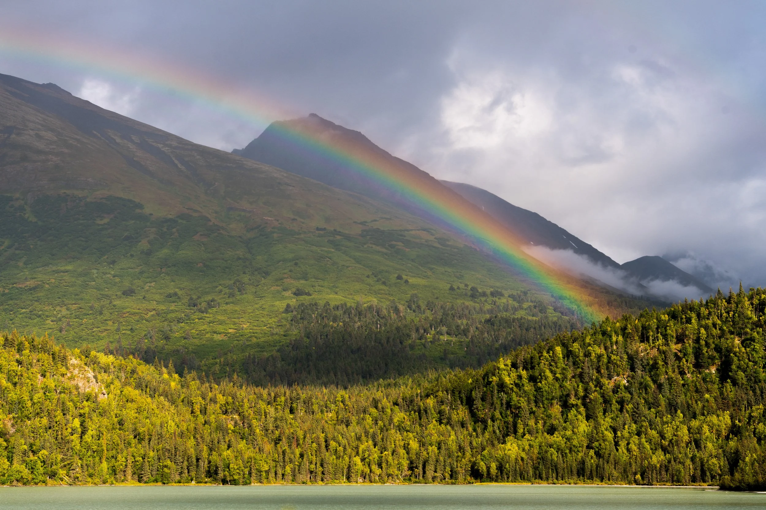 Alaska — full rainbow arcing over a mountain valley, fall foliage in yellow and green below, low clouds.