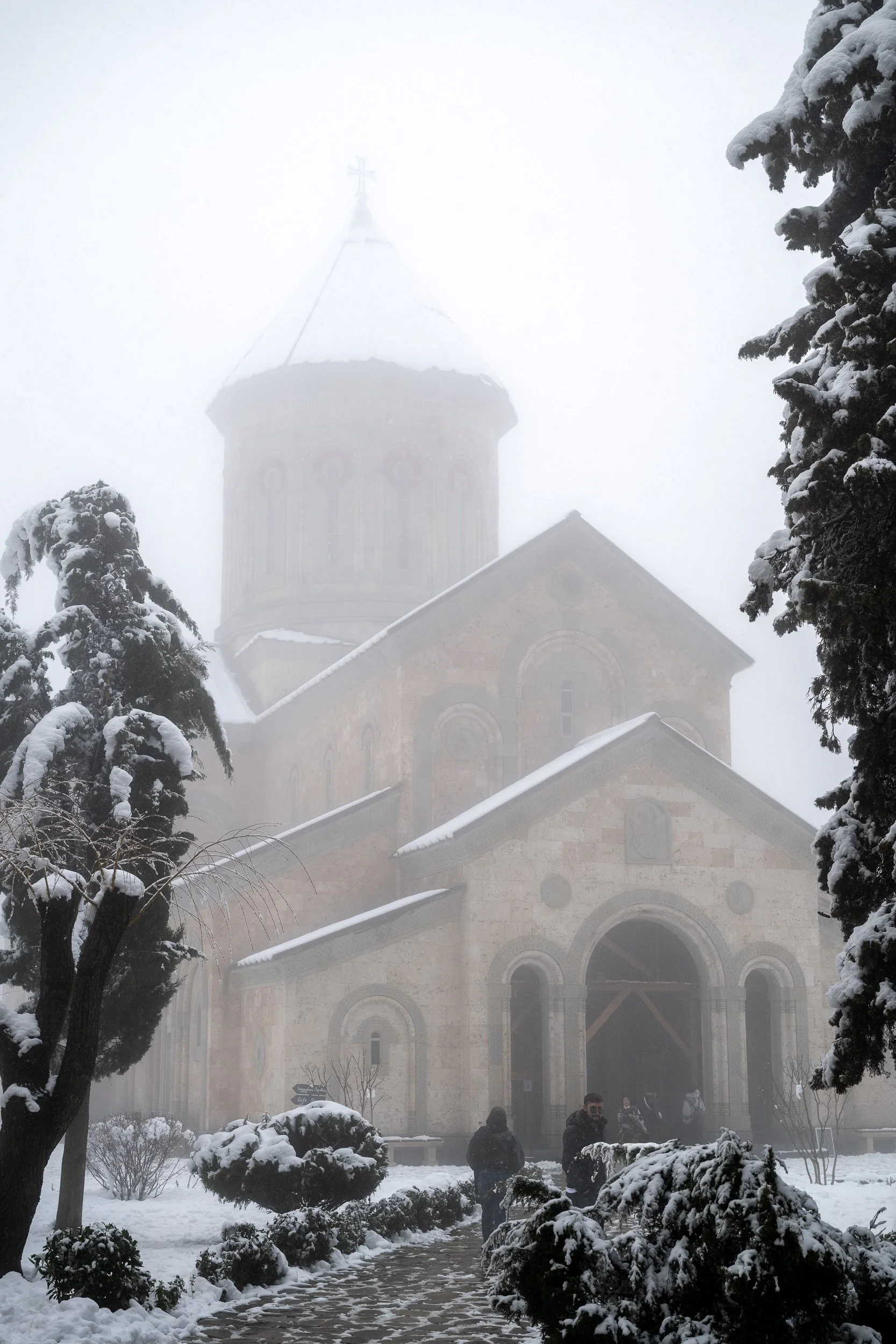 An ancient stone church emerges through snowfall and mist, its silhouette softened by the surrounding winter landscape.