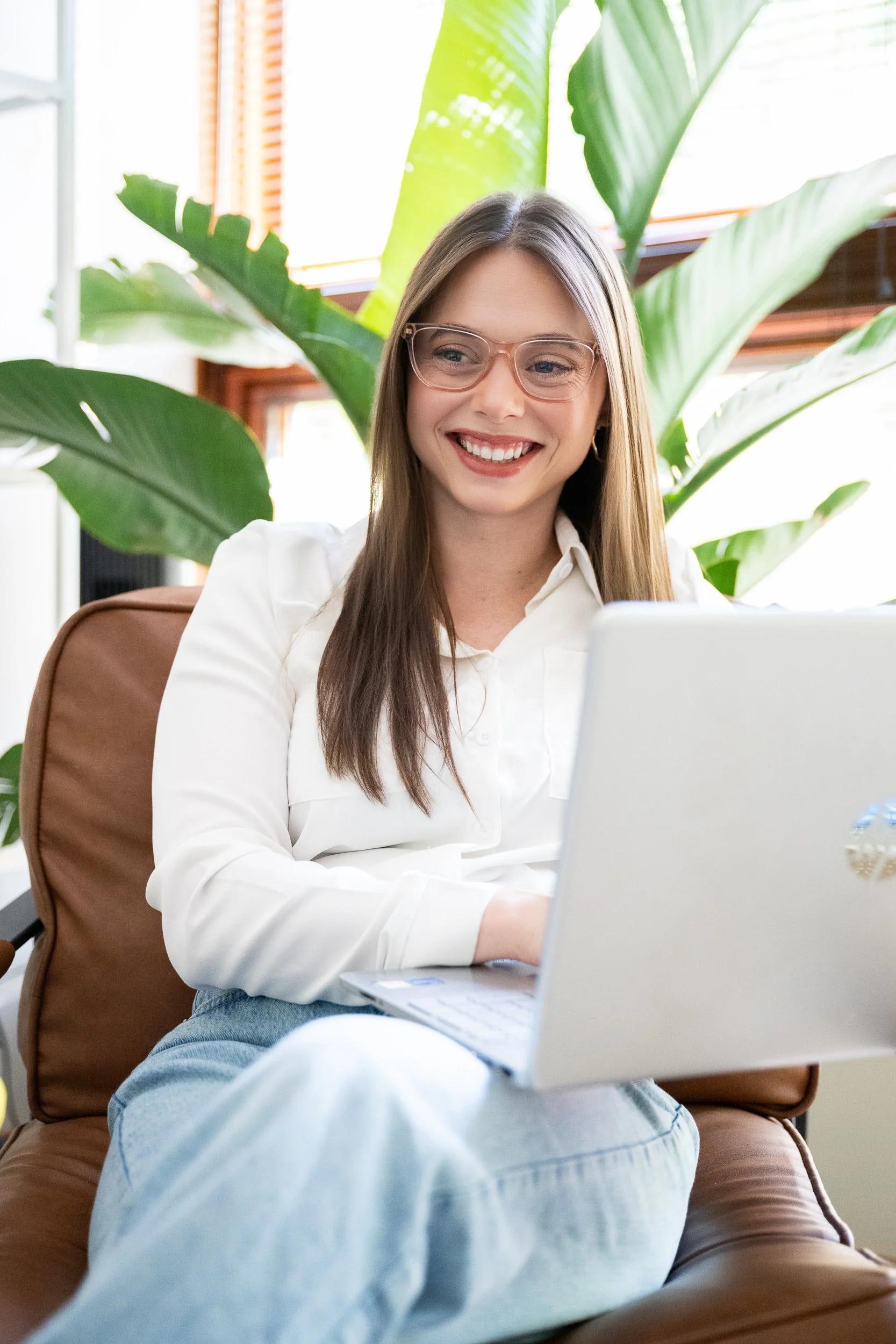 Woman with glasses smiling while working on a laptop in a plant-filled interior space