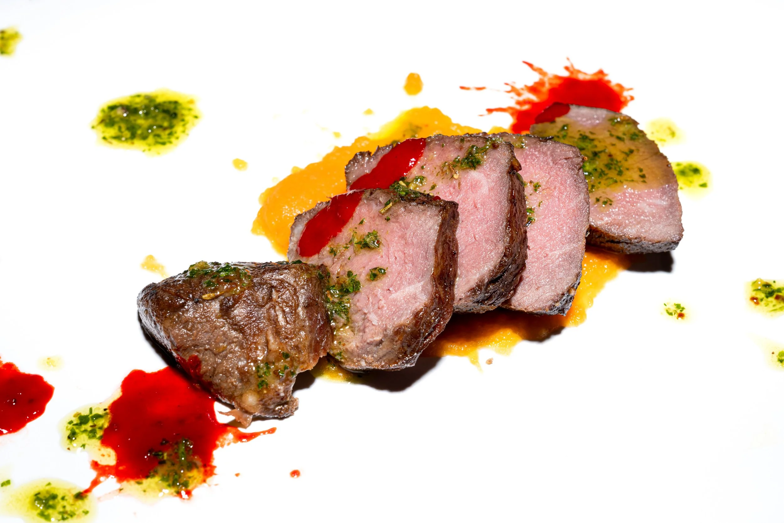 Sliced lamb with chimichurri sauce dots on white plate, fine dining restaurant food photography
