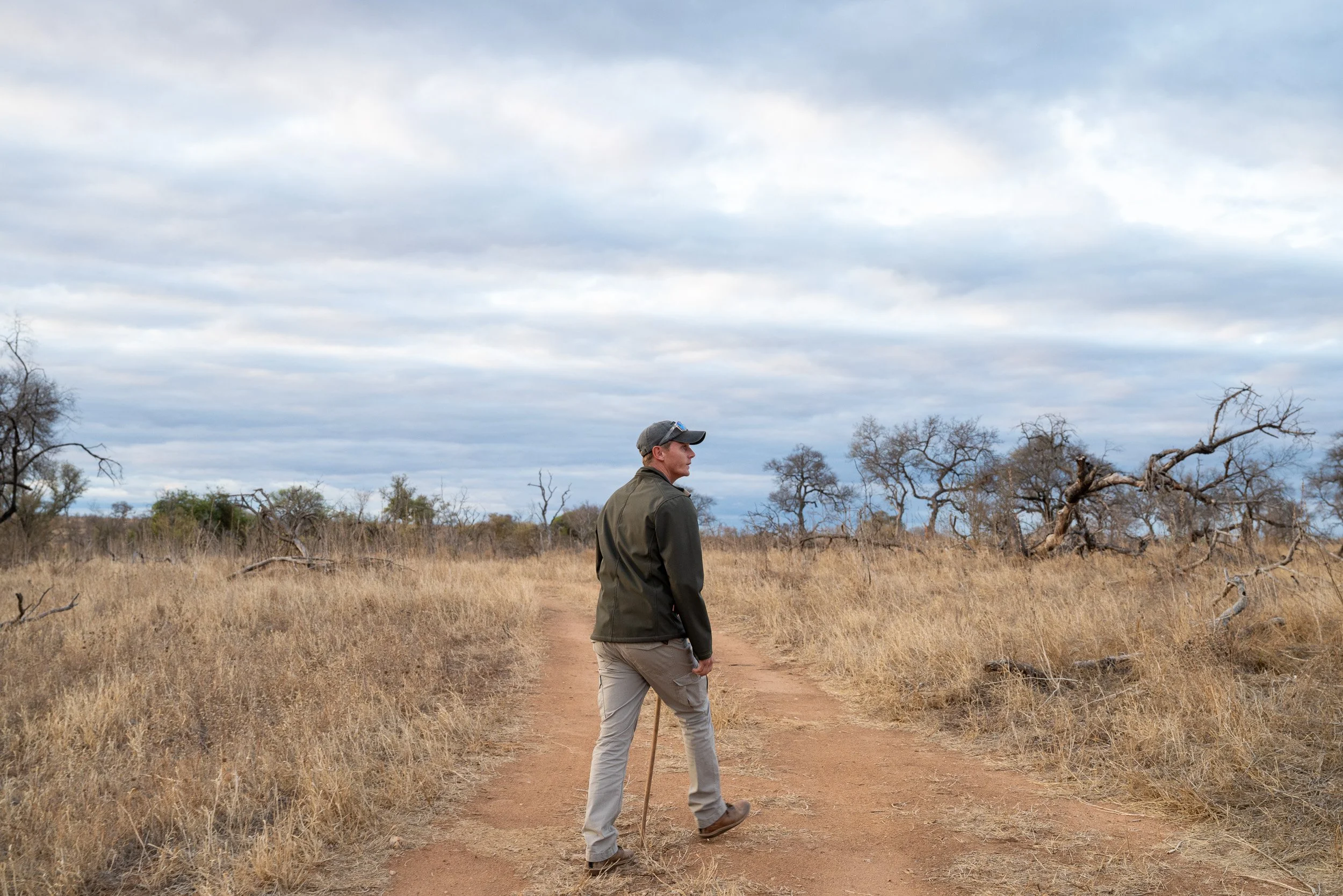 Safari guide walking along a dirt road through dry grassland.