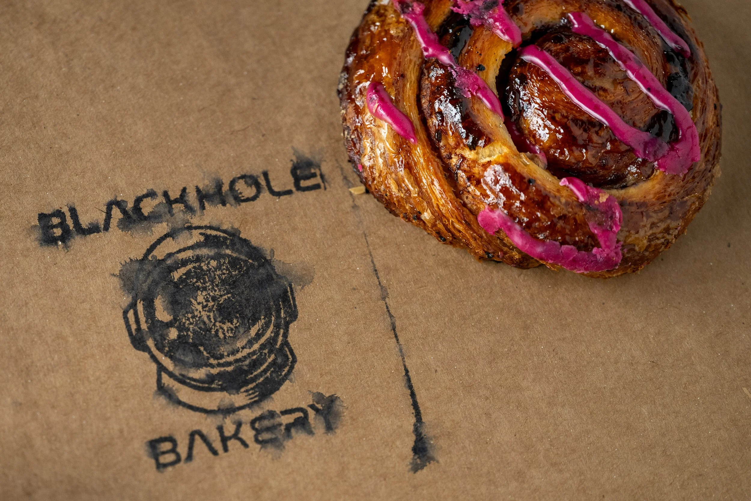 Cinnamon roll with magenta glaze beside Blackhole Bakery stamp on kraft paper, Kansas City bakery brand photography
