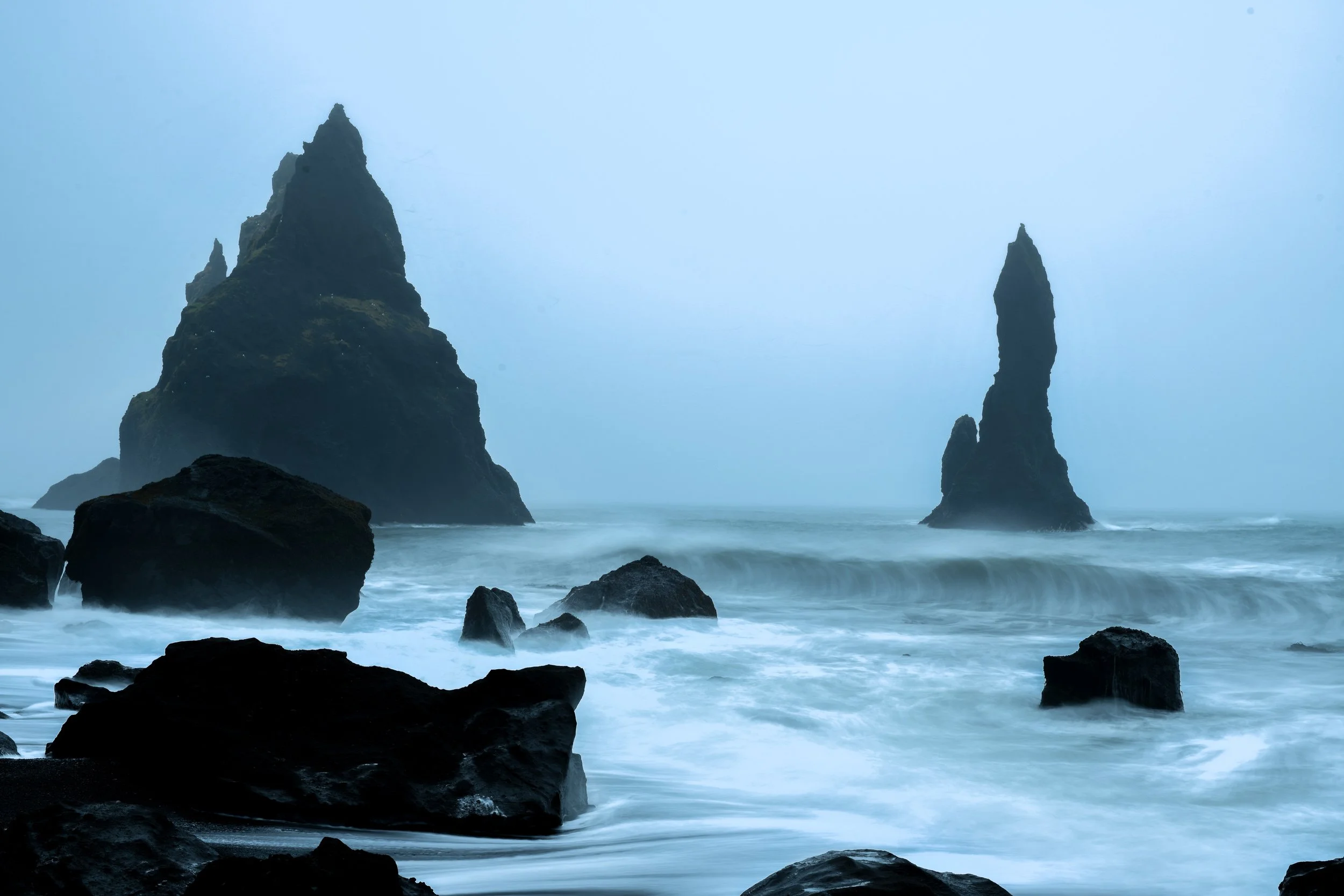 Reynisfjara black sand beach, Iceland — sea stacks rising from blue mist, long exposure waves washing over black rock.