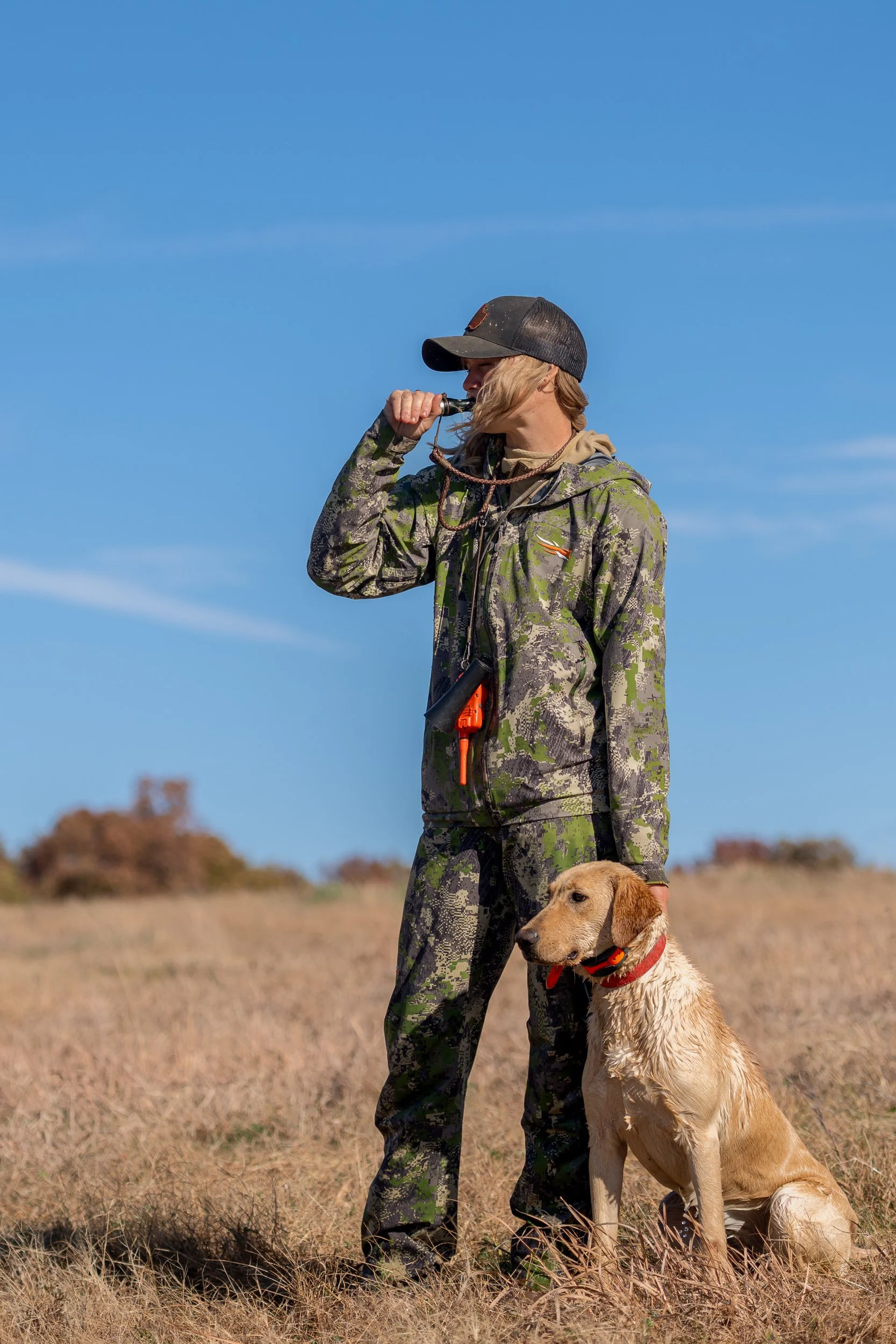 Hunter in Sitka camouflage blowing a training whistle with a yellow Labrador retriever seated at heel in an open prairie field at Habitat Flats hunting preserve in Marceline, Missouri.