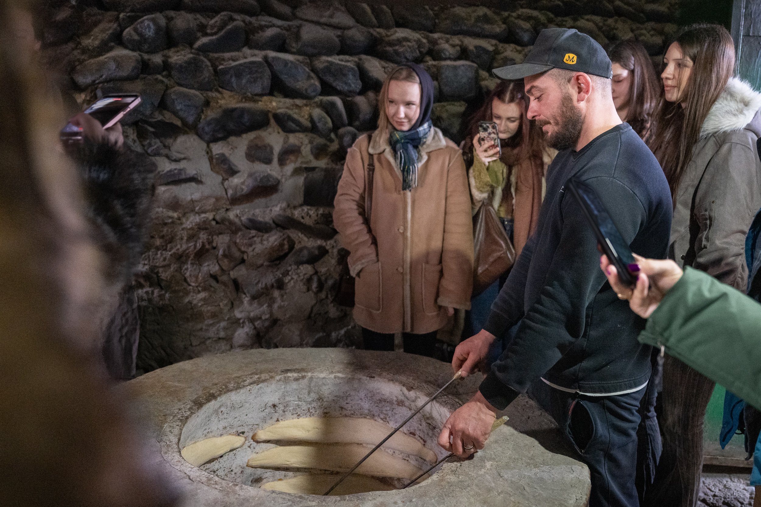 A group of women and a man gather around a large round stone oven, with some holding smartphones. They are indoors with a stone wall in the background, and the man is in the process of baking flatbread.