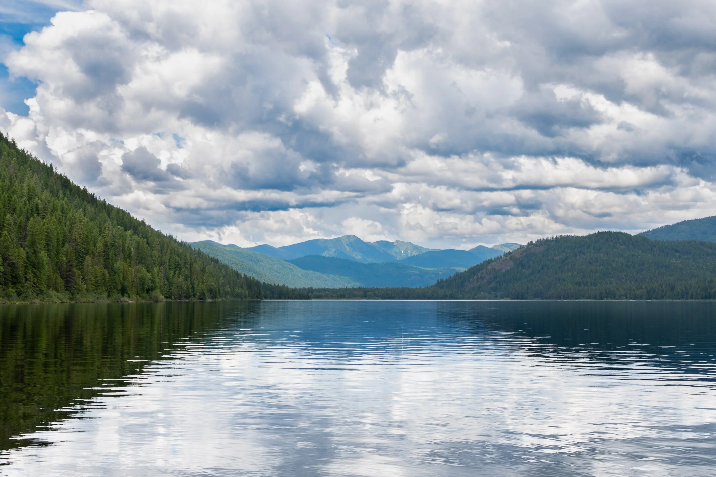 A scenic view of a large lake surrounded by forested mountains under a sky filled with cloudy weather.