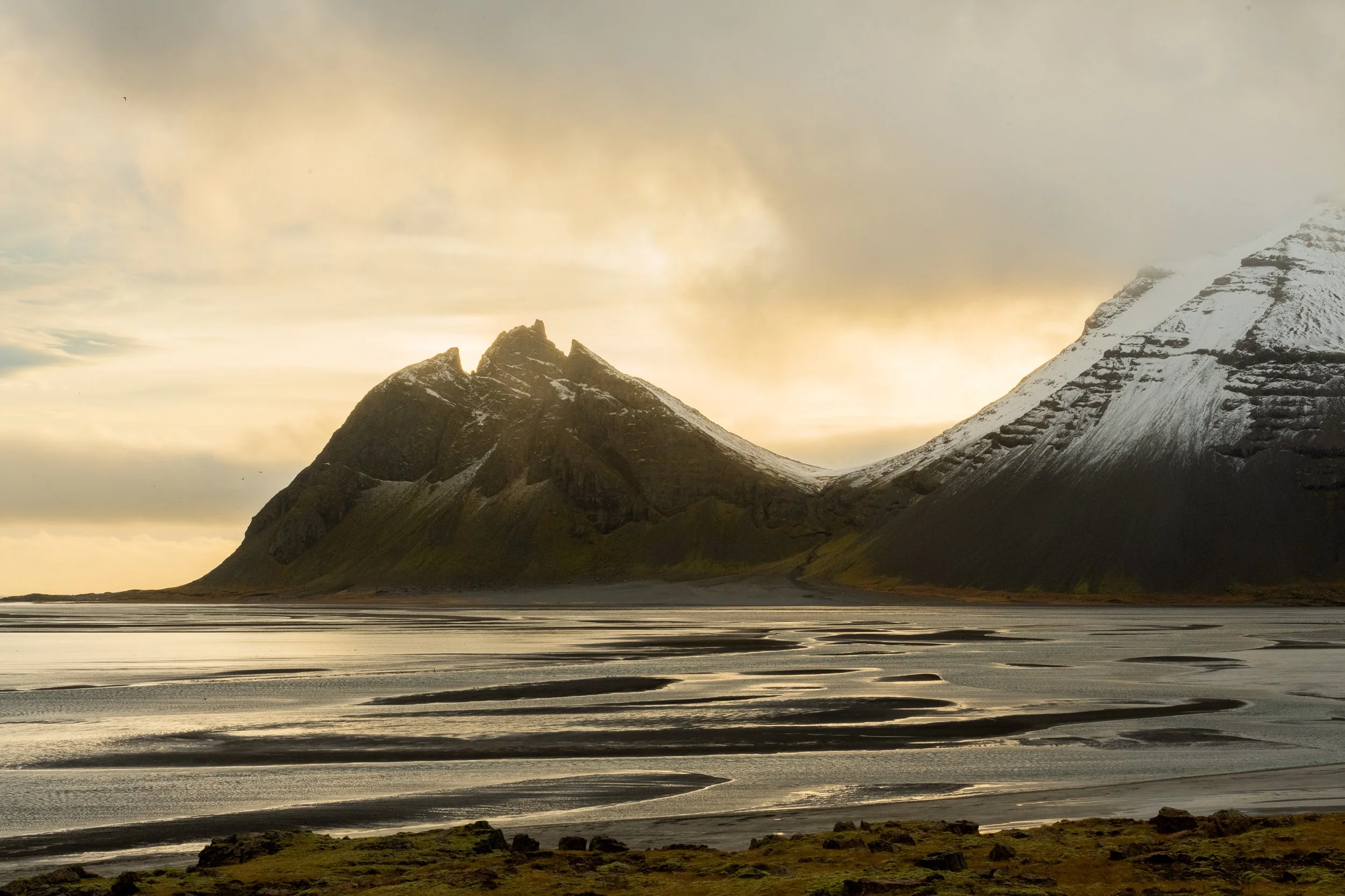 Vestrahorn mountain, Iceland — golden light raking across tidal flats below, dramatic overcast sky breaking.