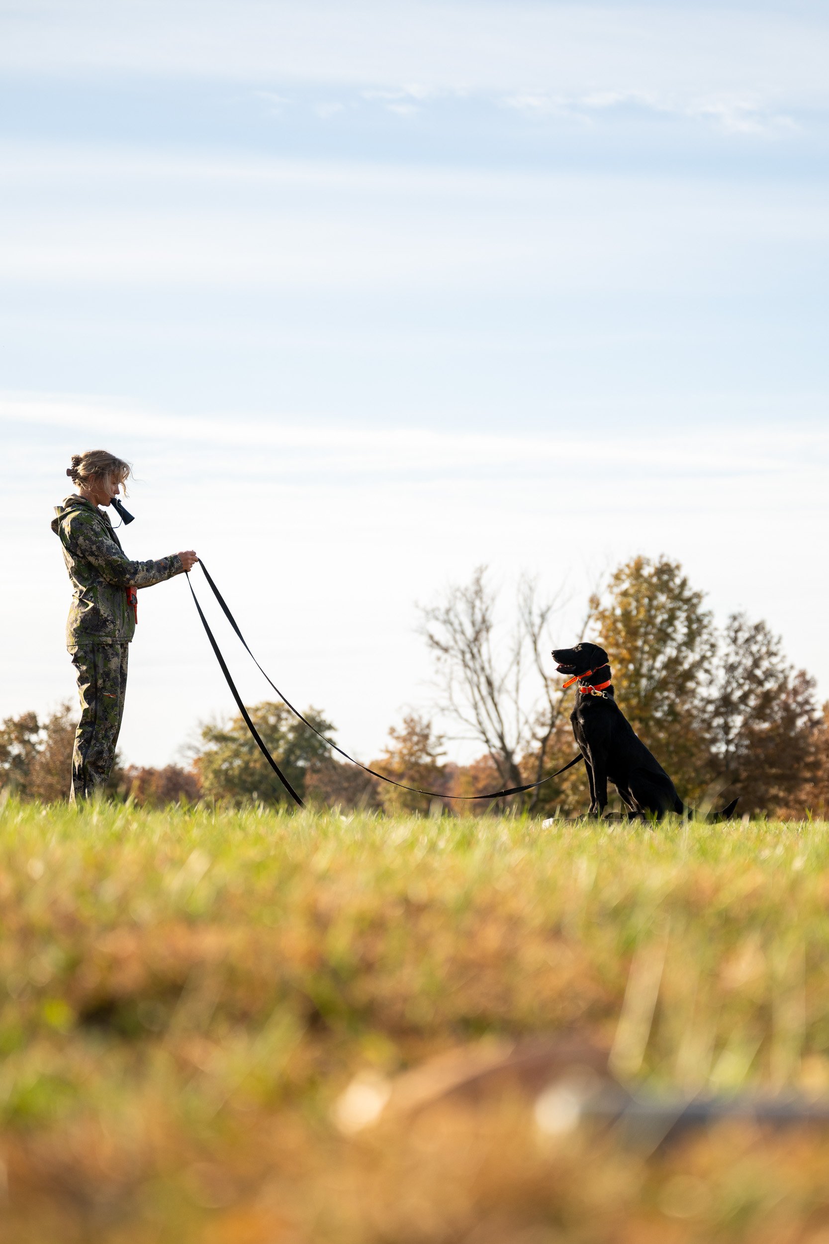 A handler works a black retriever from a distance during a marking drill in an open field.