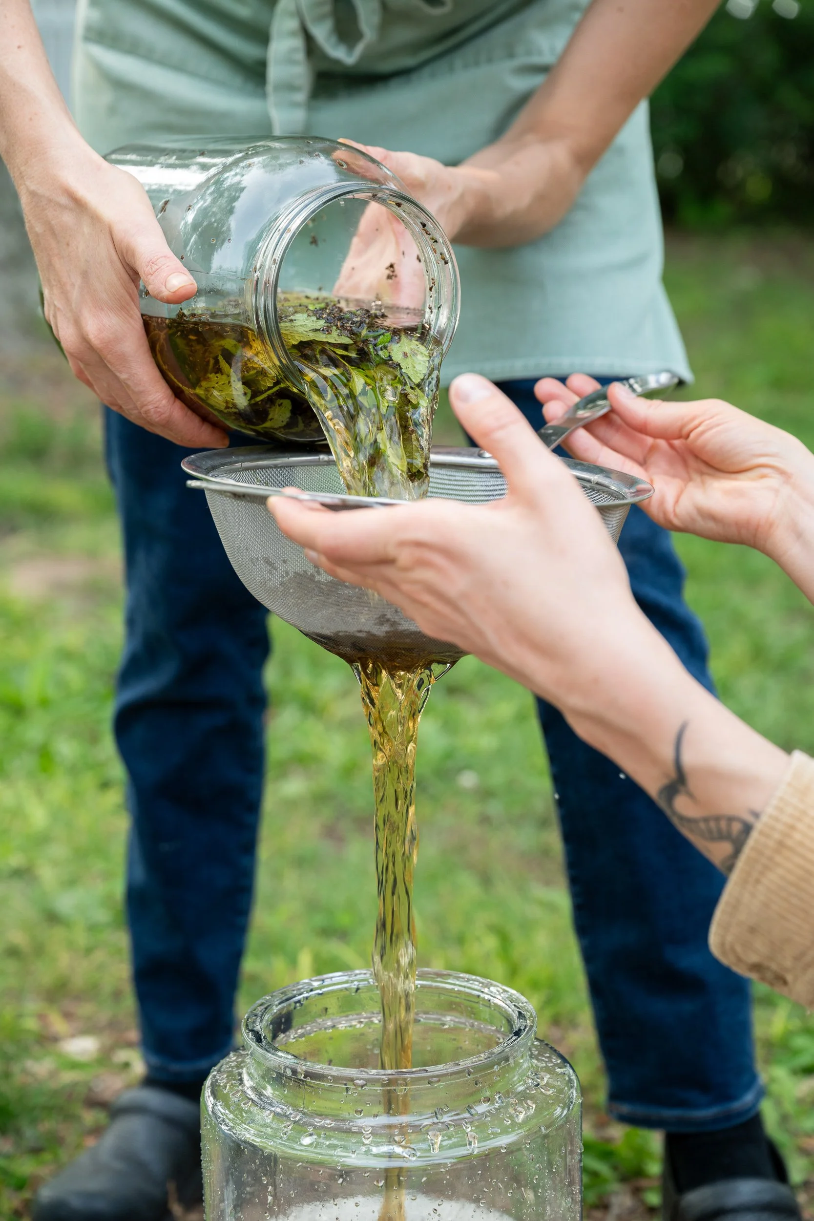 Hands straining herbal liquid through a mesh sieve into a glass jar outdoors