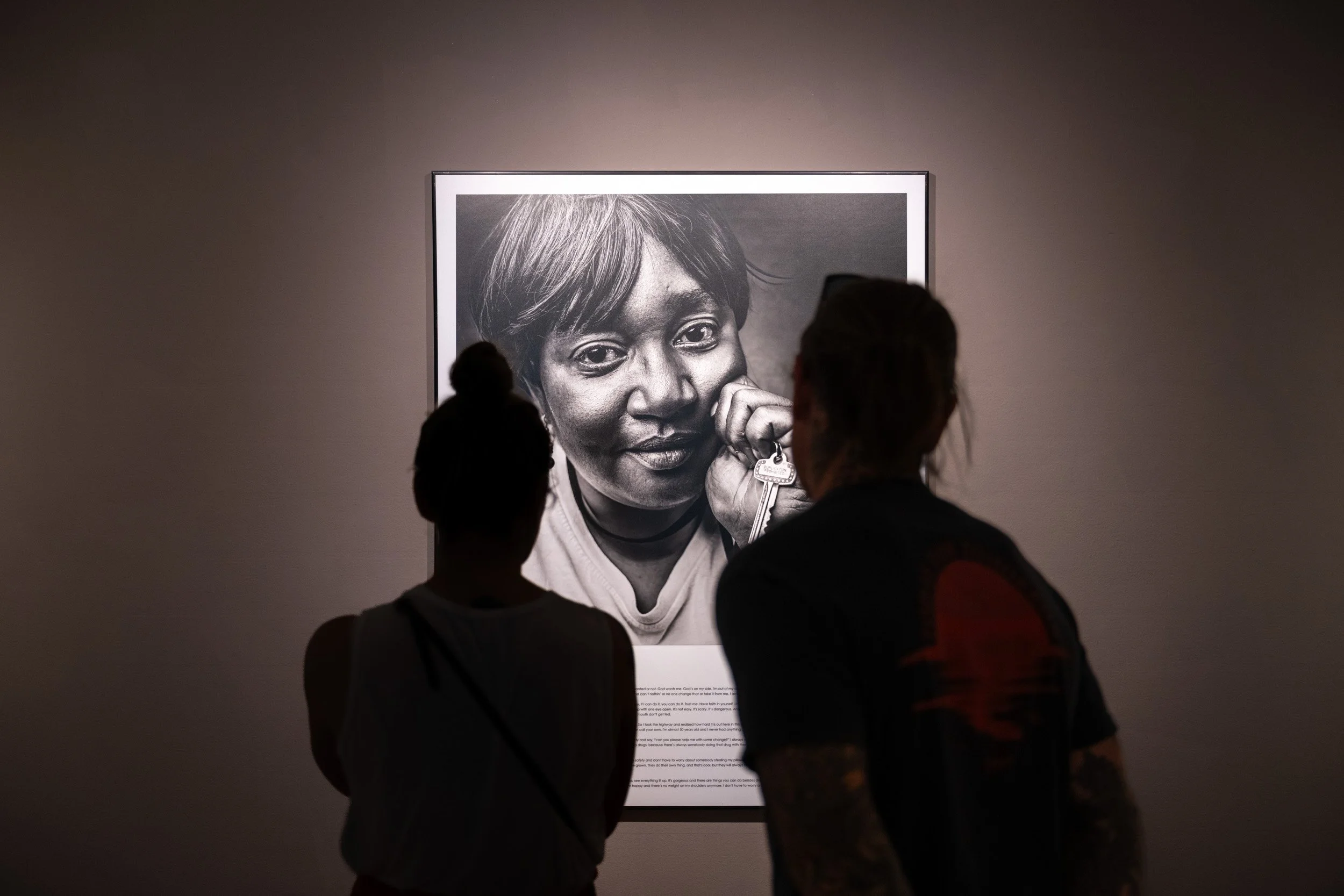 Two visitors silhouetted before a large illuminated charcoal portrait at a gallery exhibition