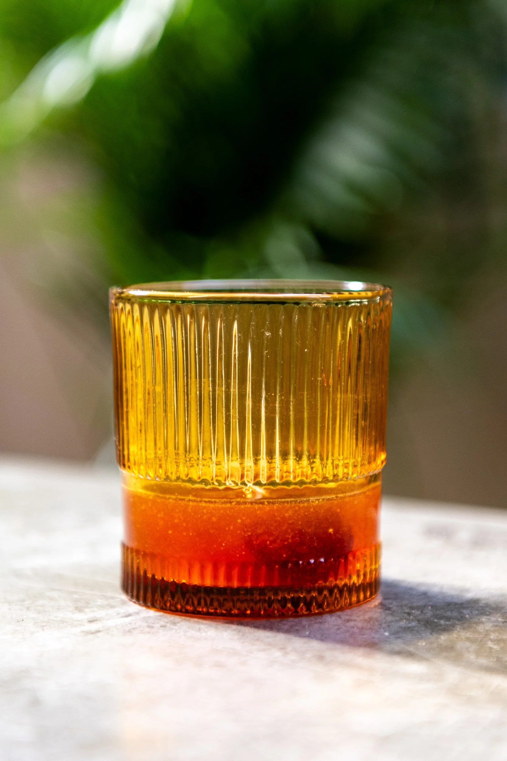 Amber cocktail in ribbed vintage glass with tropical greenery, hospitality beverage photography
