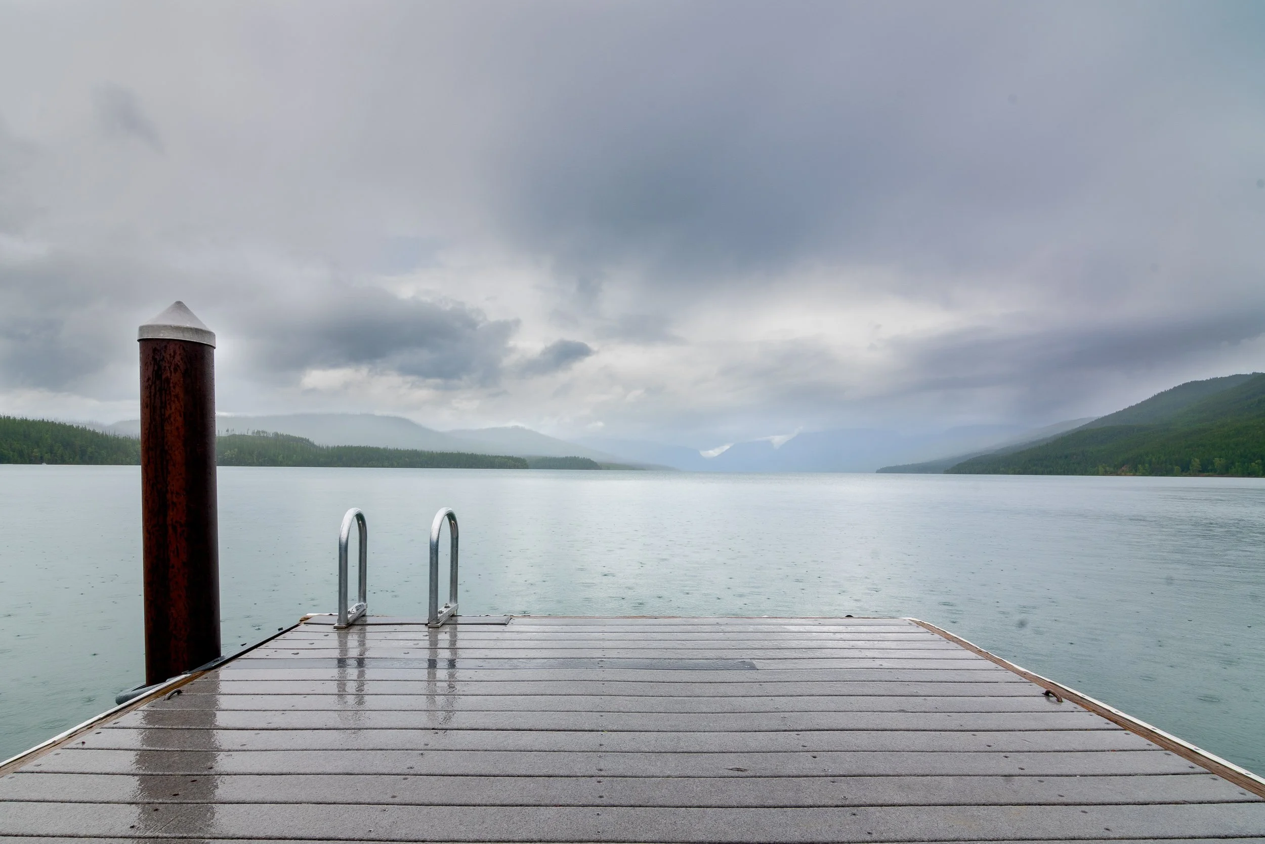  Dock extending into Lake McDonald in Glacier National Park, Montana, overcast sky, forested mountains along the shoreline.