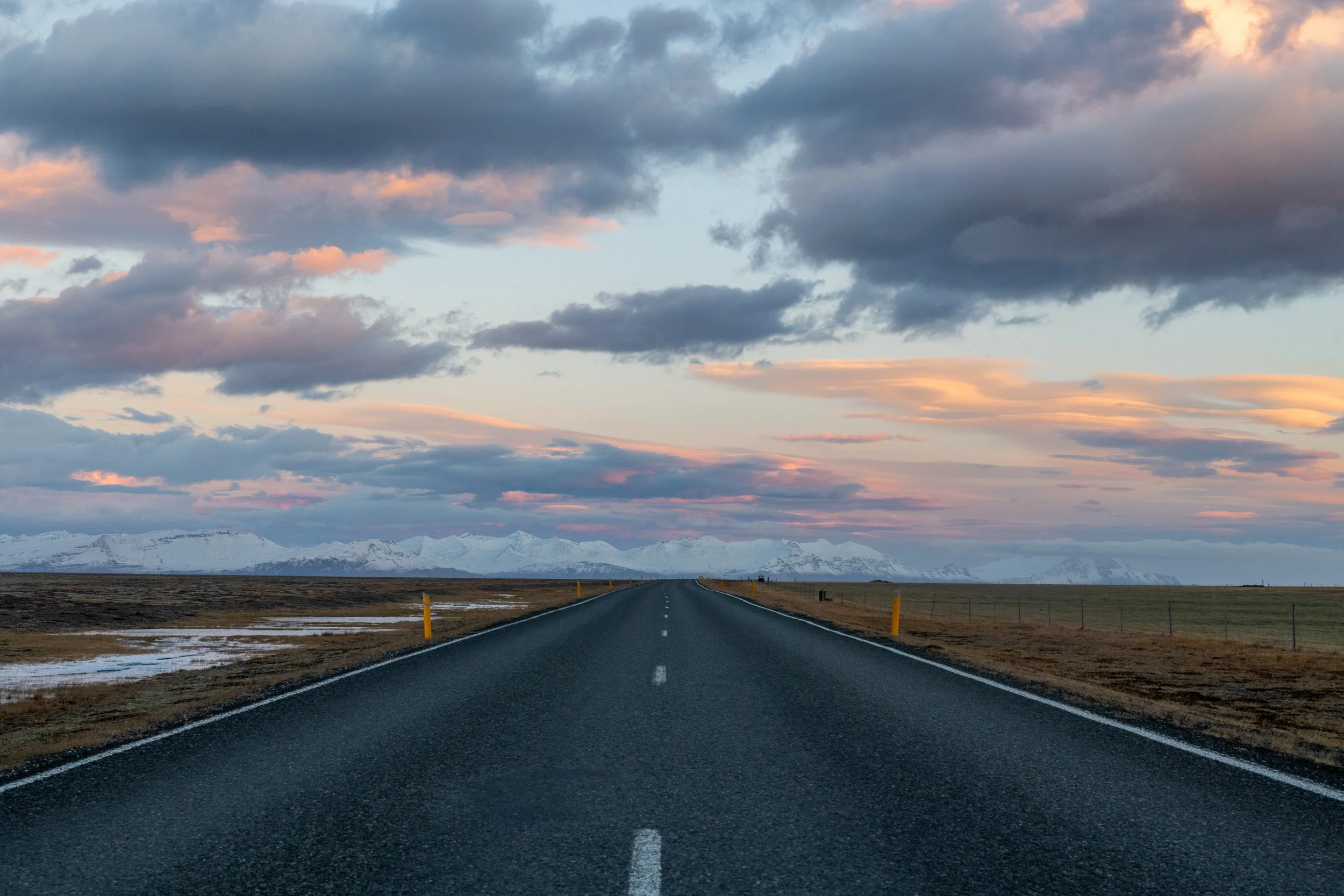  Straight road across an Icelandic plain at dusk, pink and blue cloudscape above, mountains in the distance.